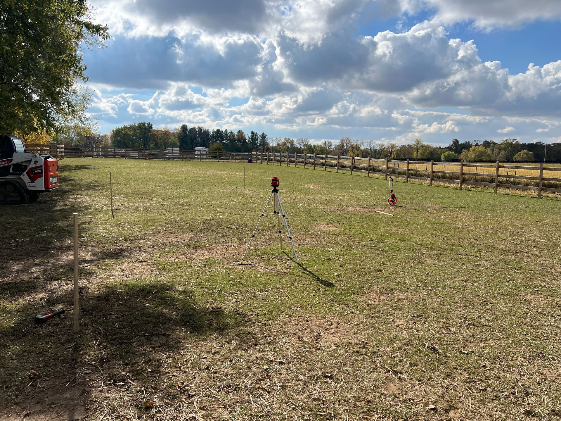 A surveying laser level on a tripod stands in a grassy field with a fence in the background and a piece of equipment nearby.