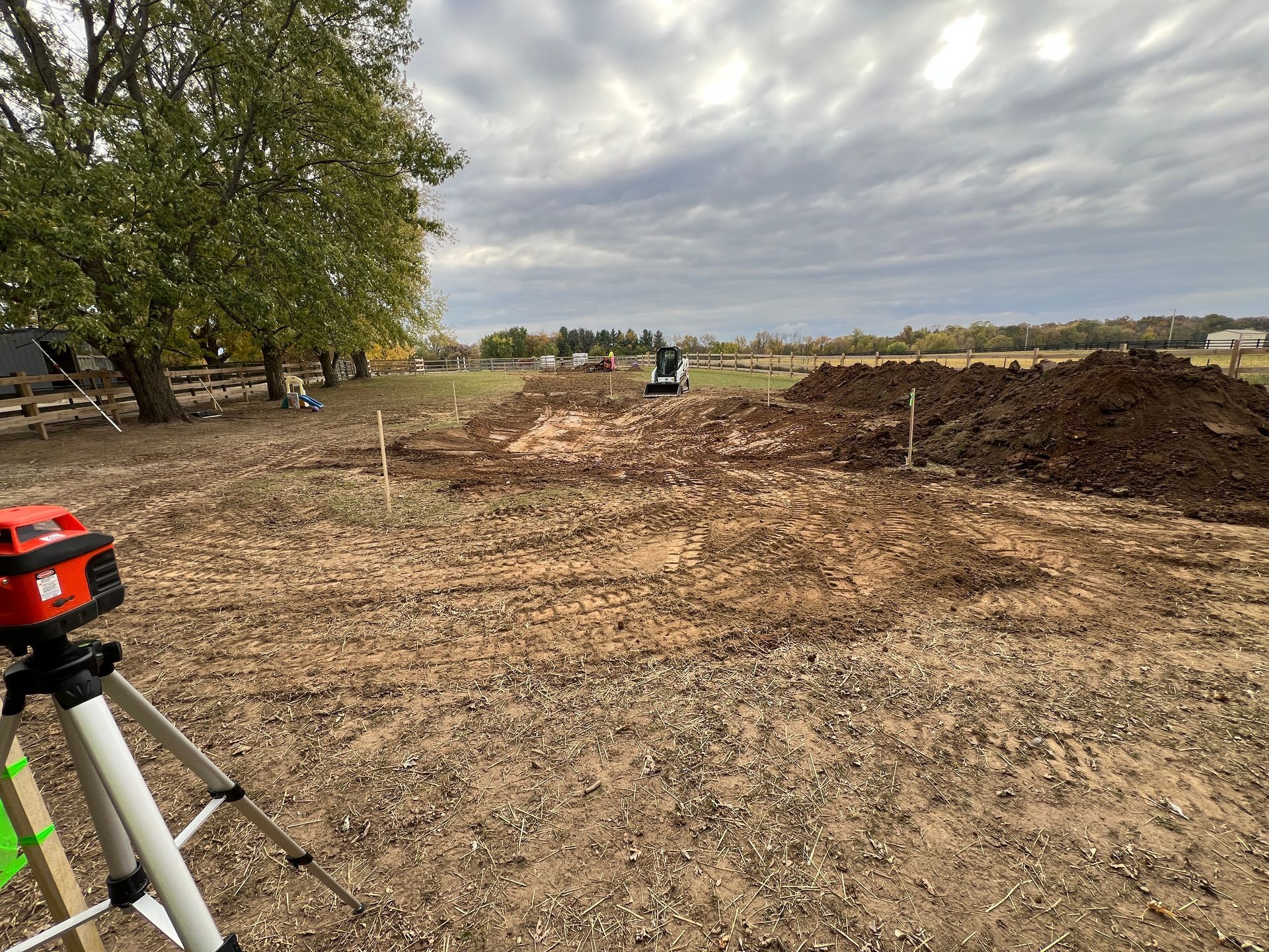 A laser level on a tripod faces an outdoor construction site with piles of dirt and a small excavator in the distance.