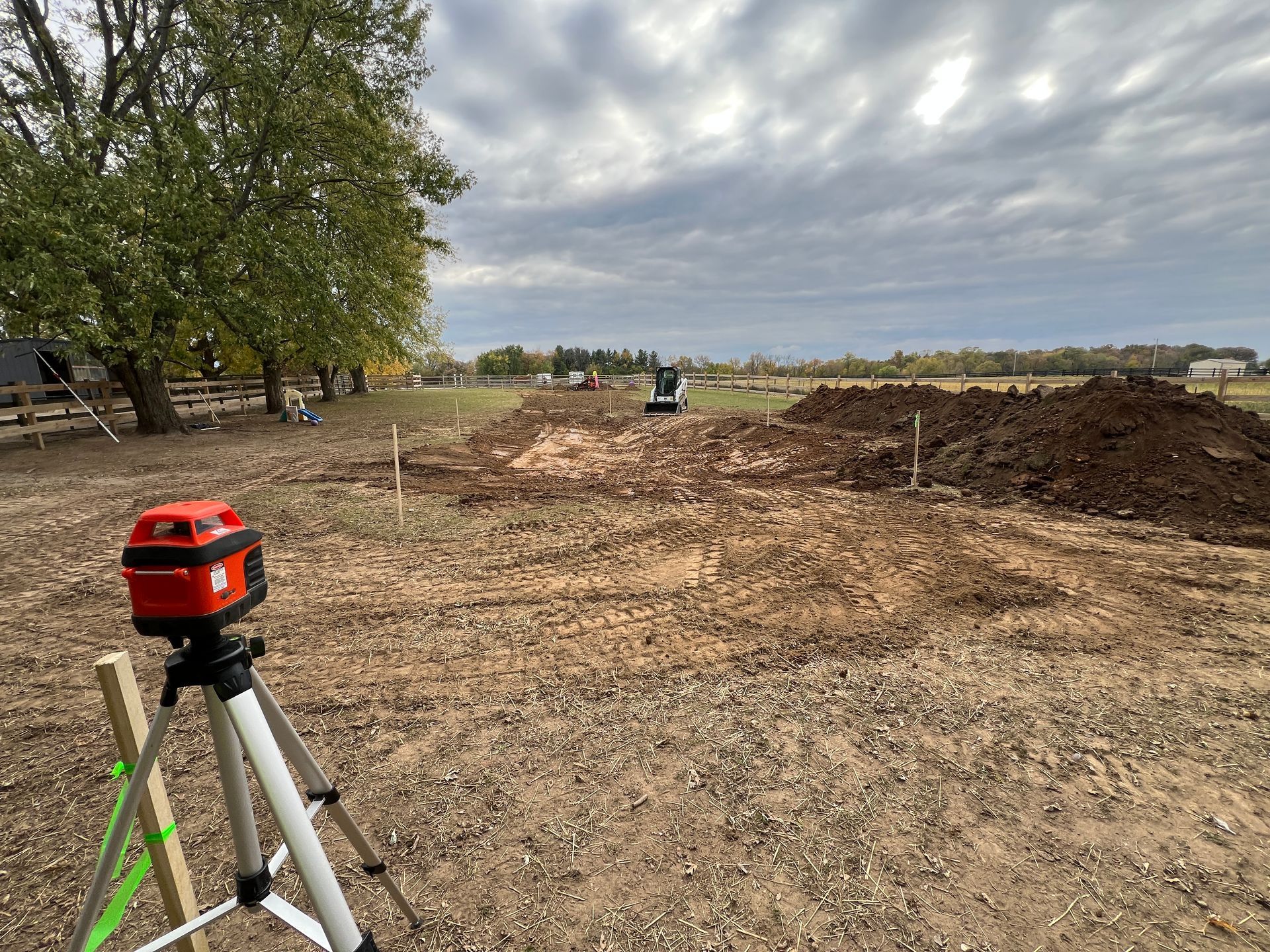A red laser level on a tripod stands in a field with excavated earth and construction equipment in the background.