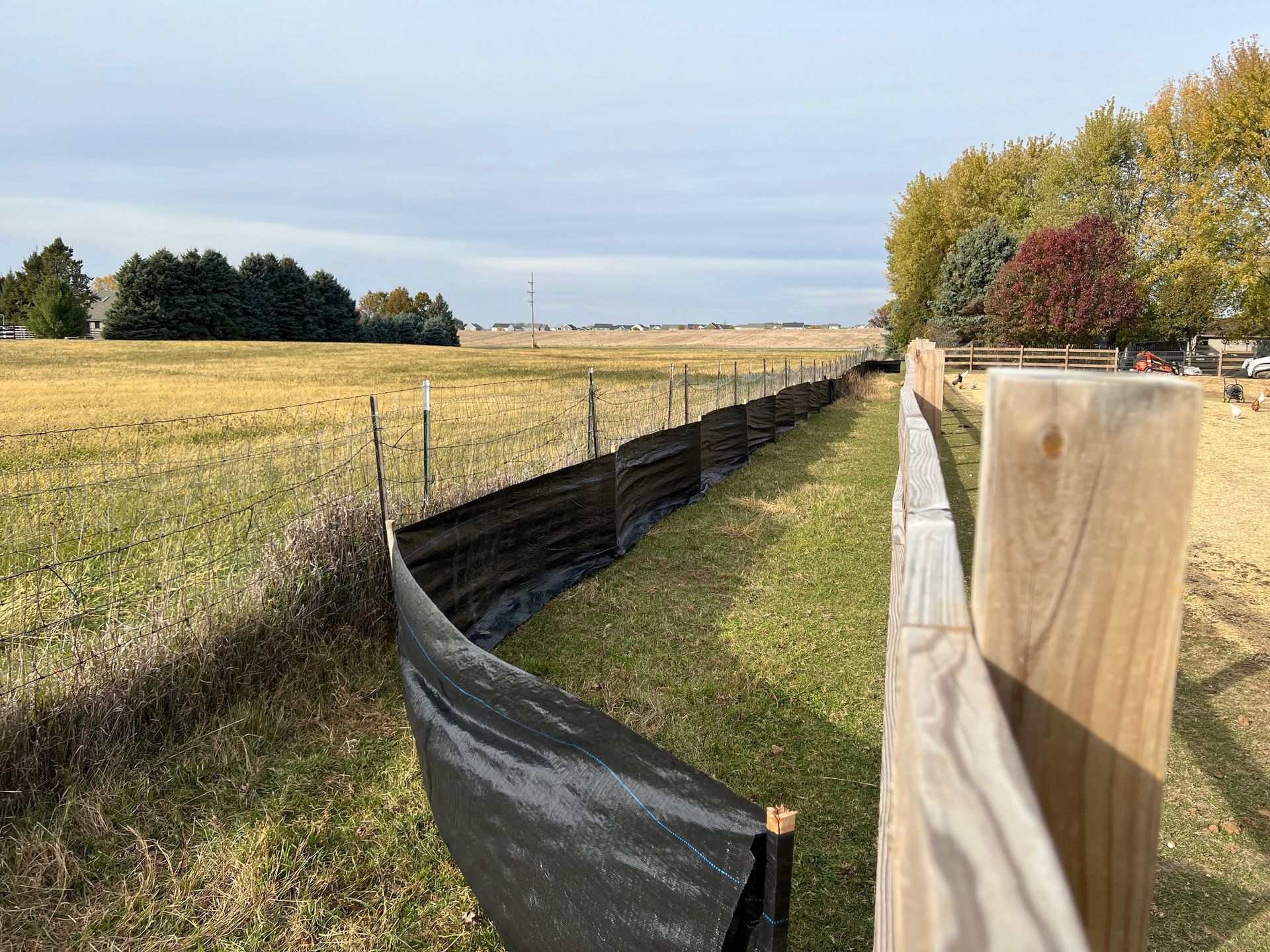 A long black silt fence follows a wooden post fence line through a grassy field toward distant trees under a bright sky.