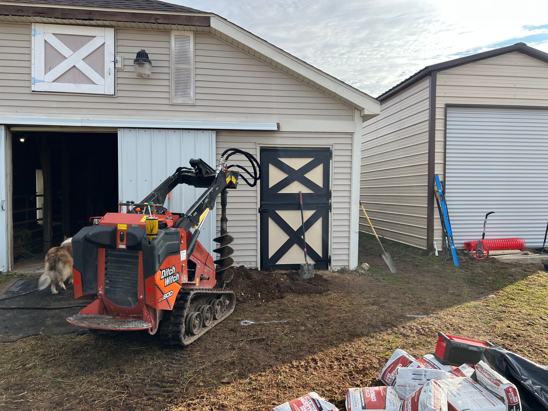 An orange skid steer with an auger attachment parked outside a barn, with a small dog and a pile of bags nearby.