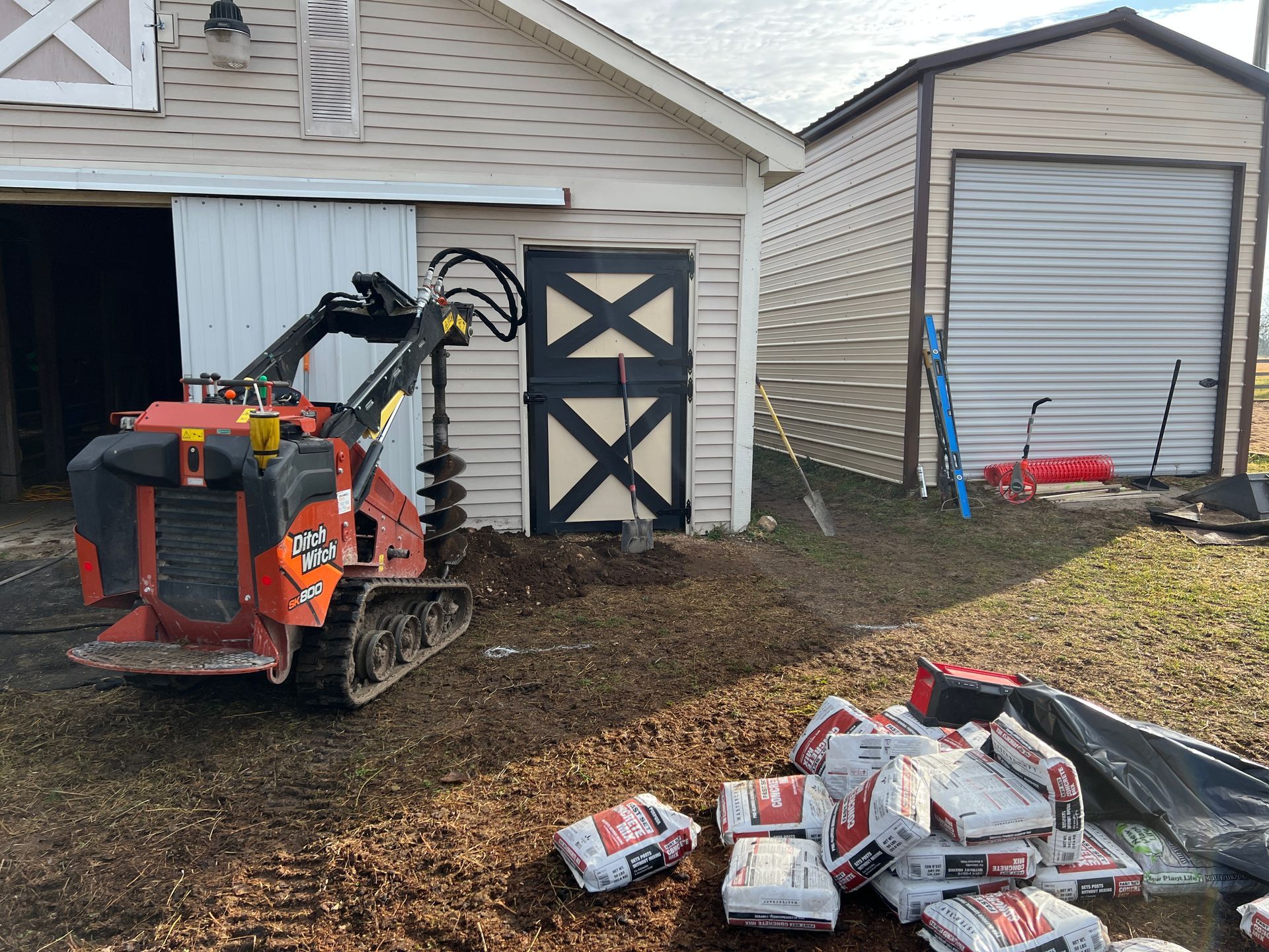 An orange skid steer with an auger attachment sits in front of a barn next to a pile of bagged concrete.