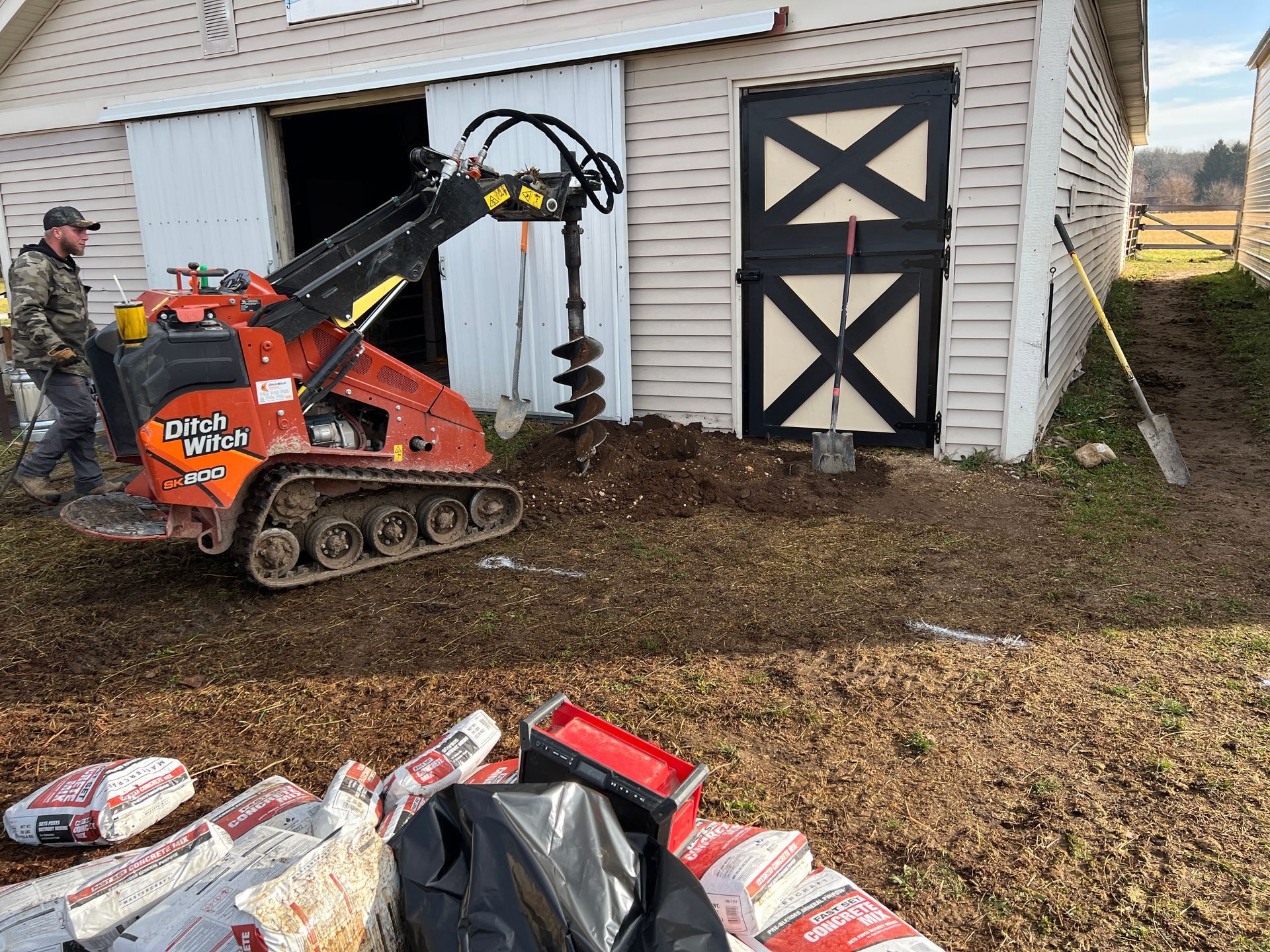 A person operates an orange compact track loader with an auger attachment near a barn, with bags of supplies in the yard.