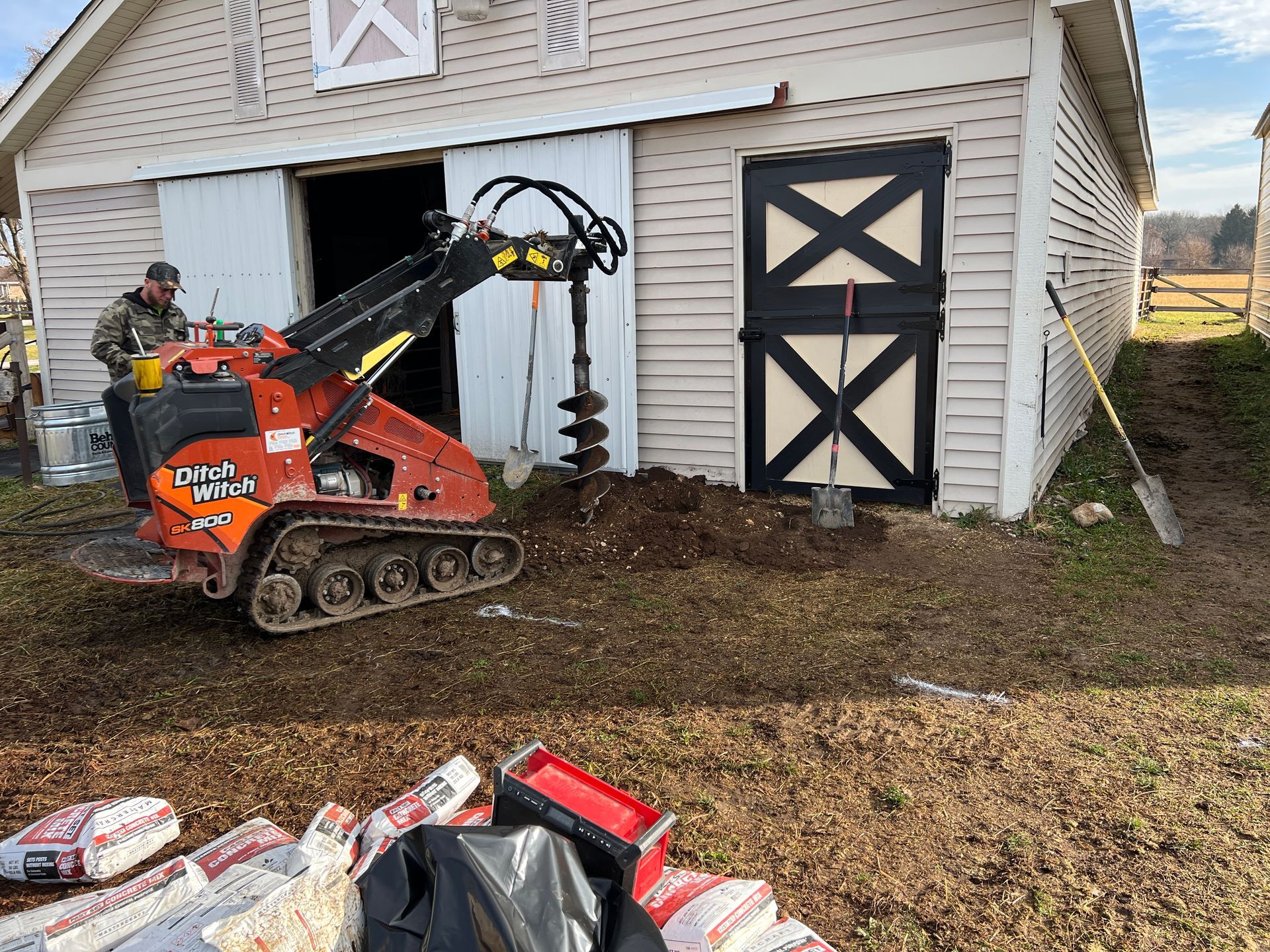 A person operates an orange compact track loader with an auger attachment outside a barn, near bags of concrete.