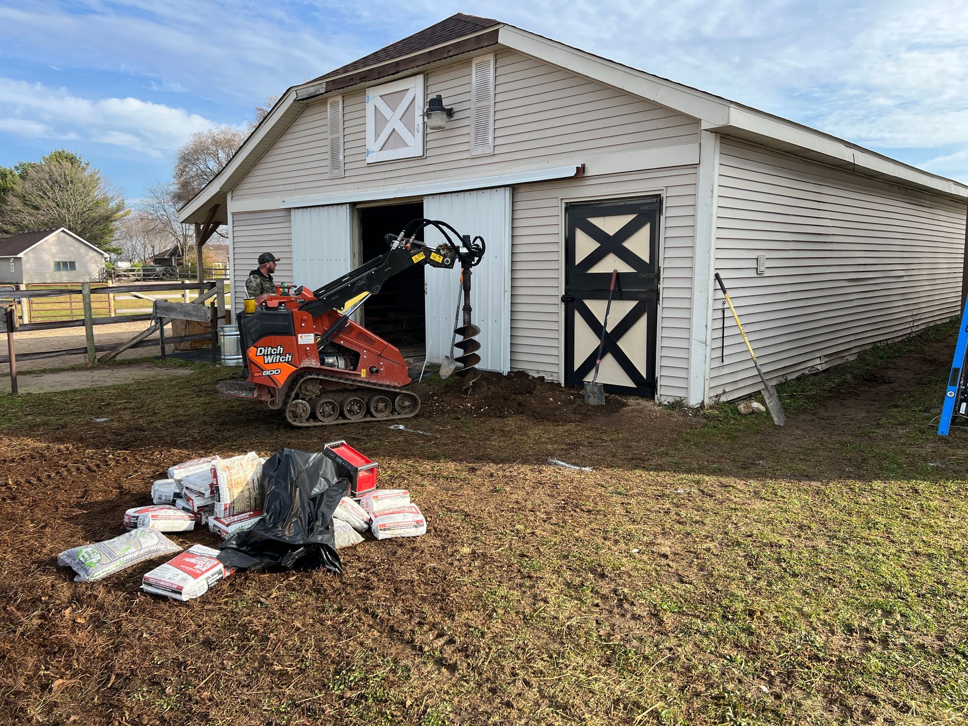 A construction worker operates an orange skid steer with an auger attachment outside a light-colored barn with sliding doors.