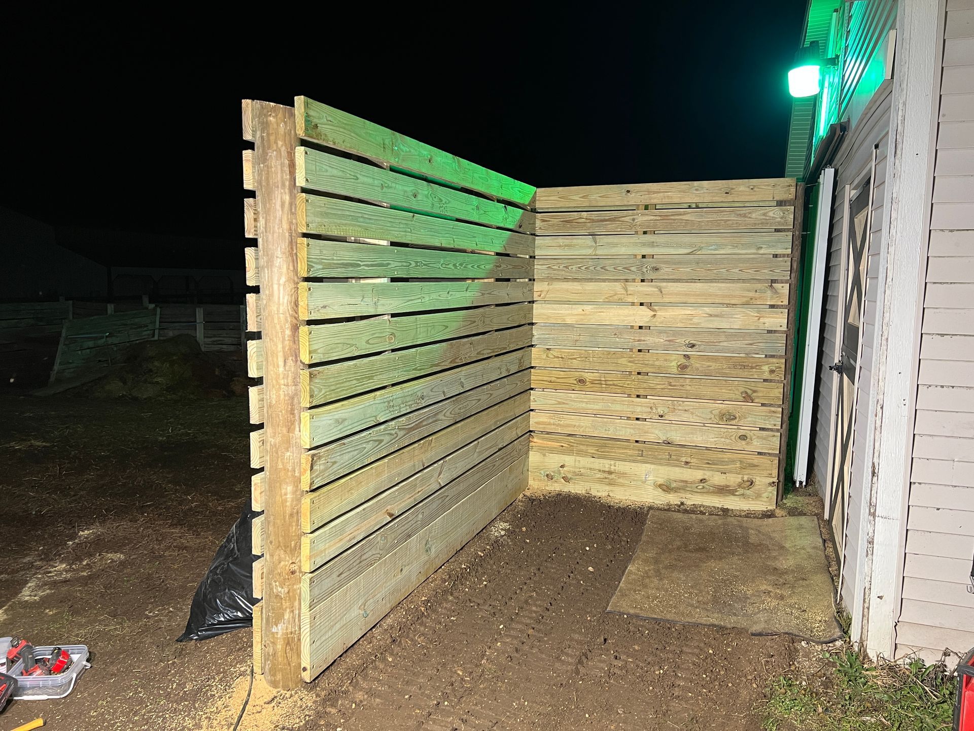 A newly constructed wooden fence with horizontal slats standing in a yard next to the exterior of a building at night.