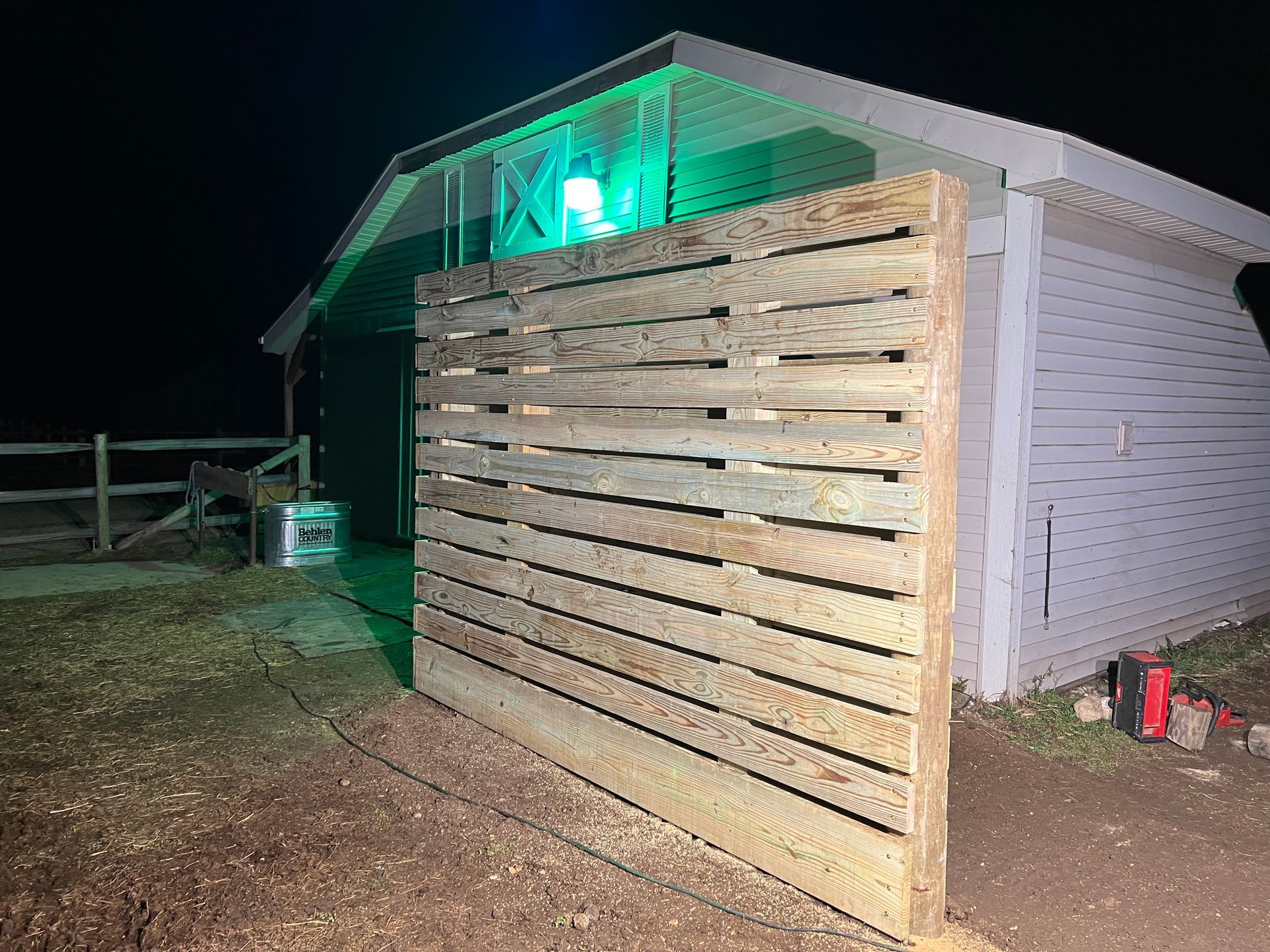 A wooden slat privacy wall stands outside a building at night, illuminated by a green light under the building's overhang.