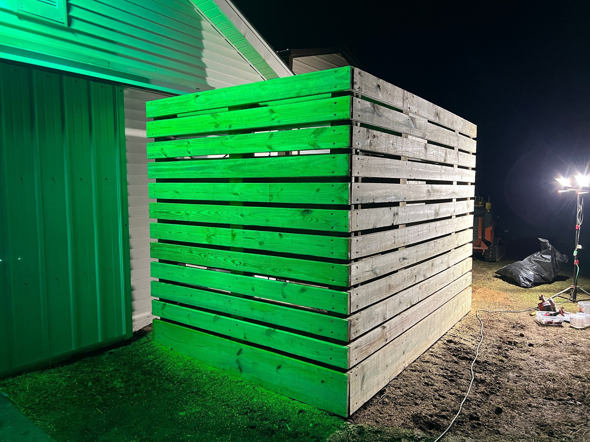 A wooden slat structure stands outside at night, illuminated by a bright green light next to a building.
