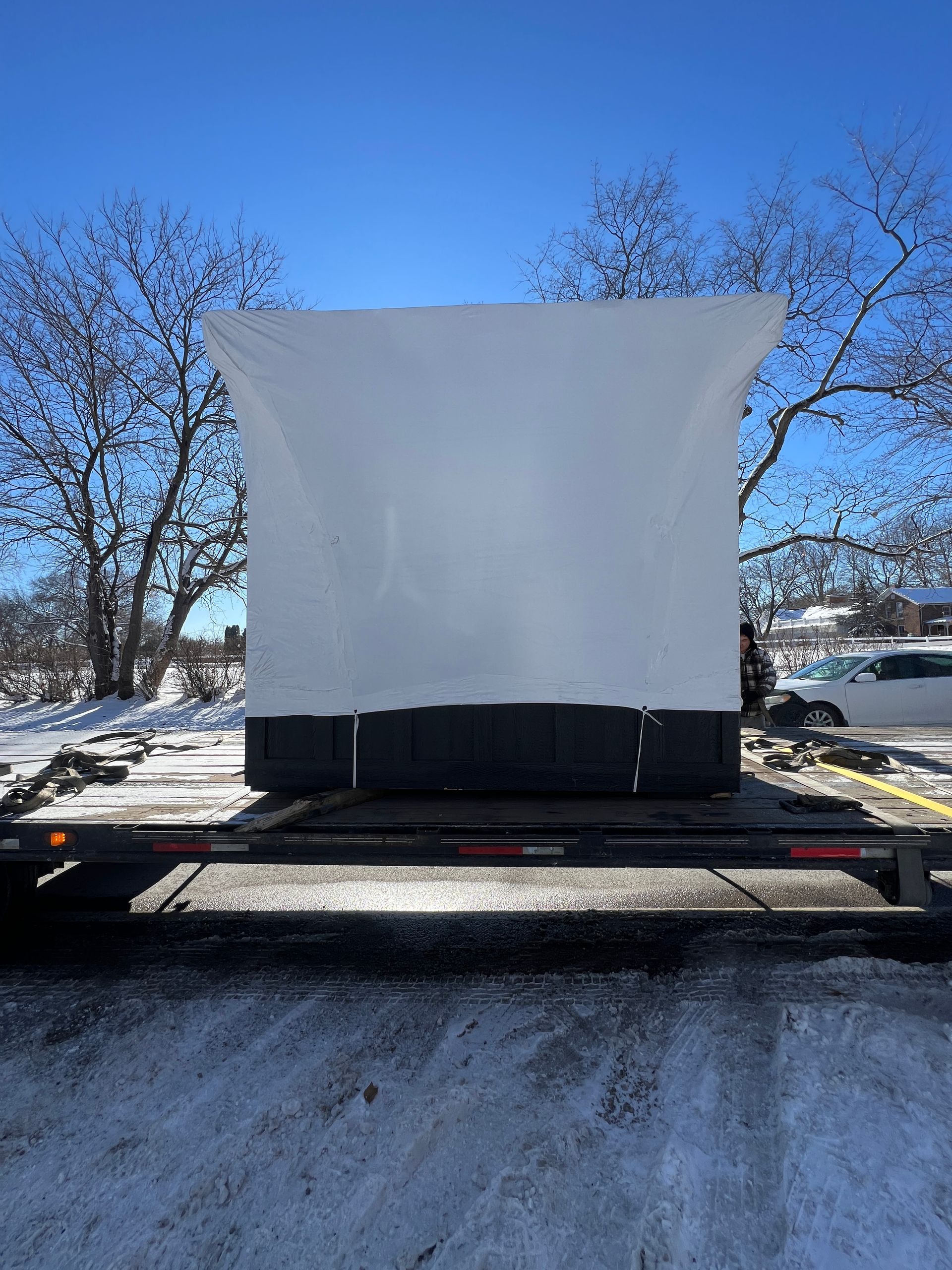 A large, white-wrapped rectangular object sits centered on a flatbed trailer in a snowy, sunny outdoor parking lot.