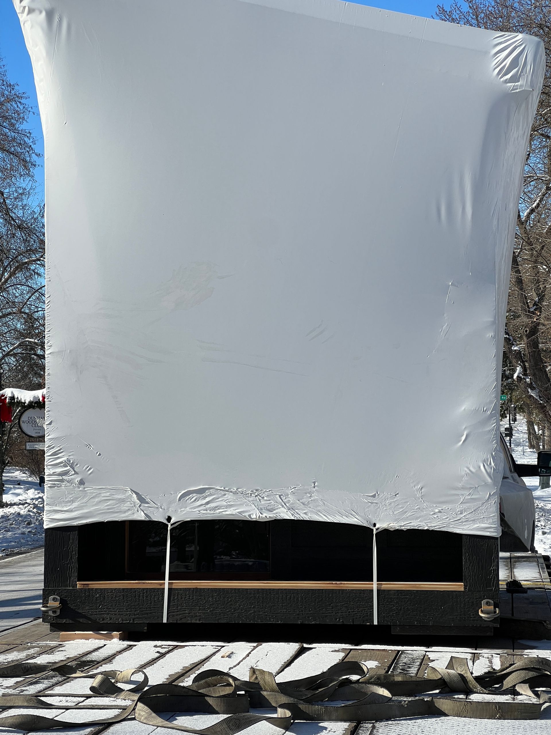 A large, white-wrapped rectangular object sits on a black pallet frame in a snowy, outdoor setting with trees.