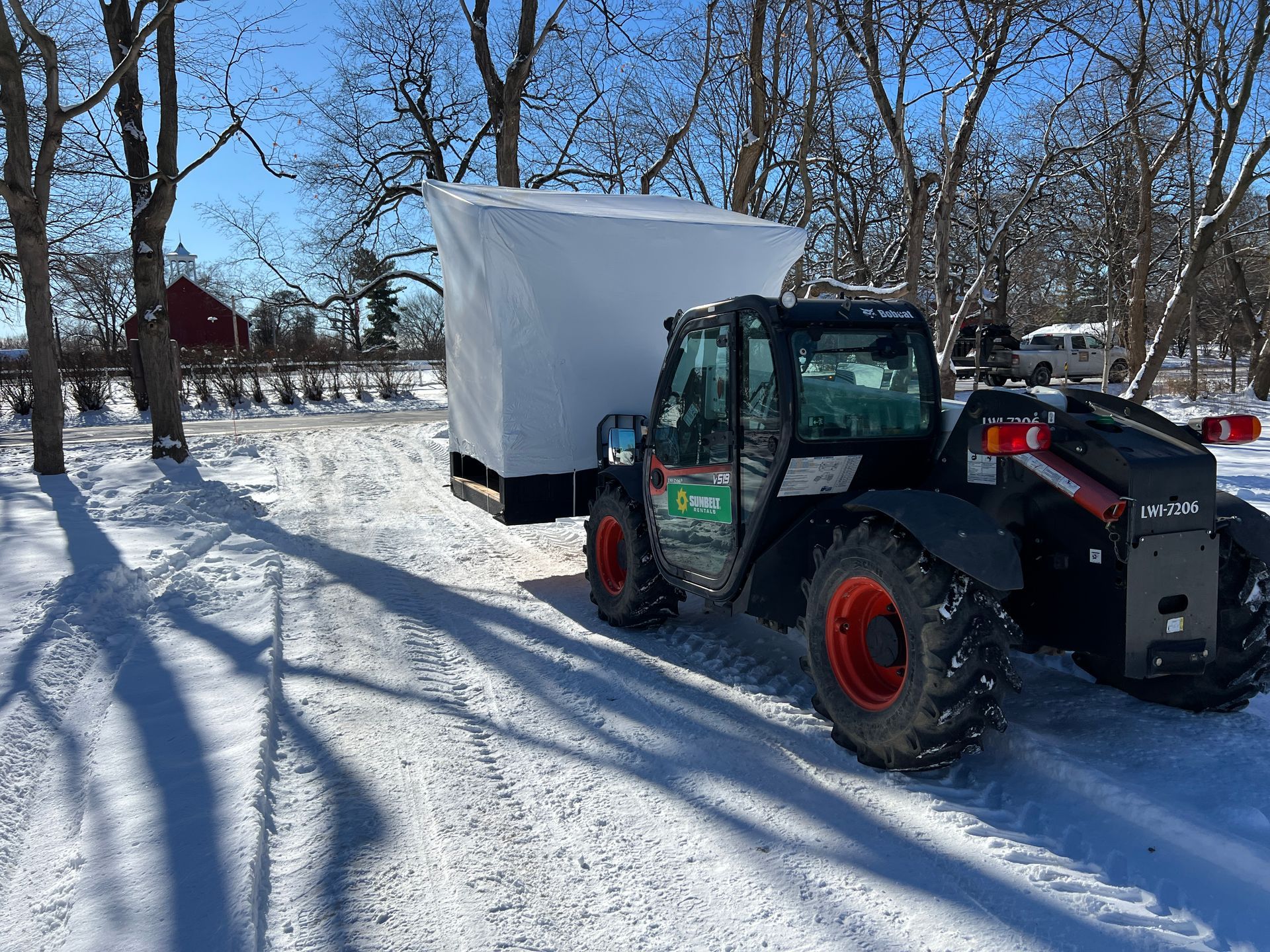 A black telehandler carries a large, white-covered crate through a snow-covered field with trees and a red barn nearby.