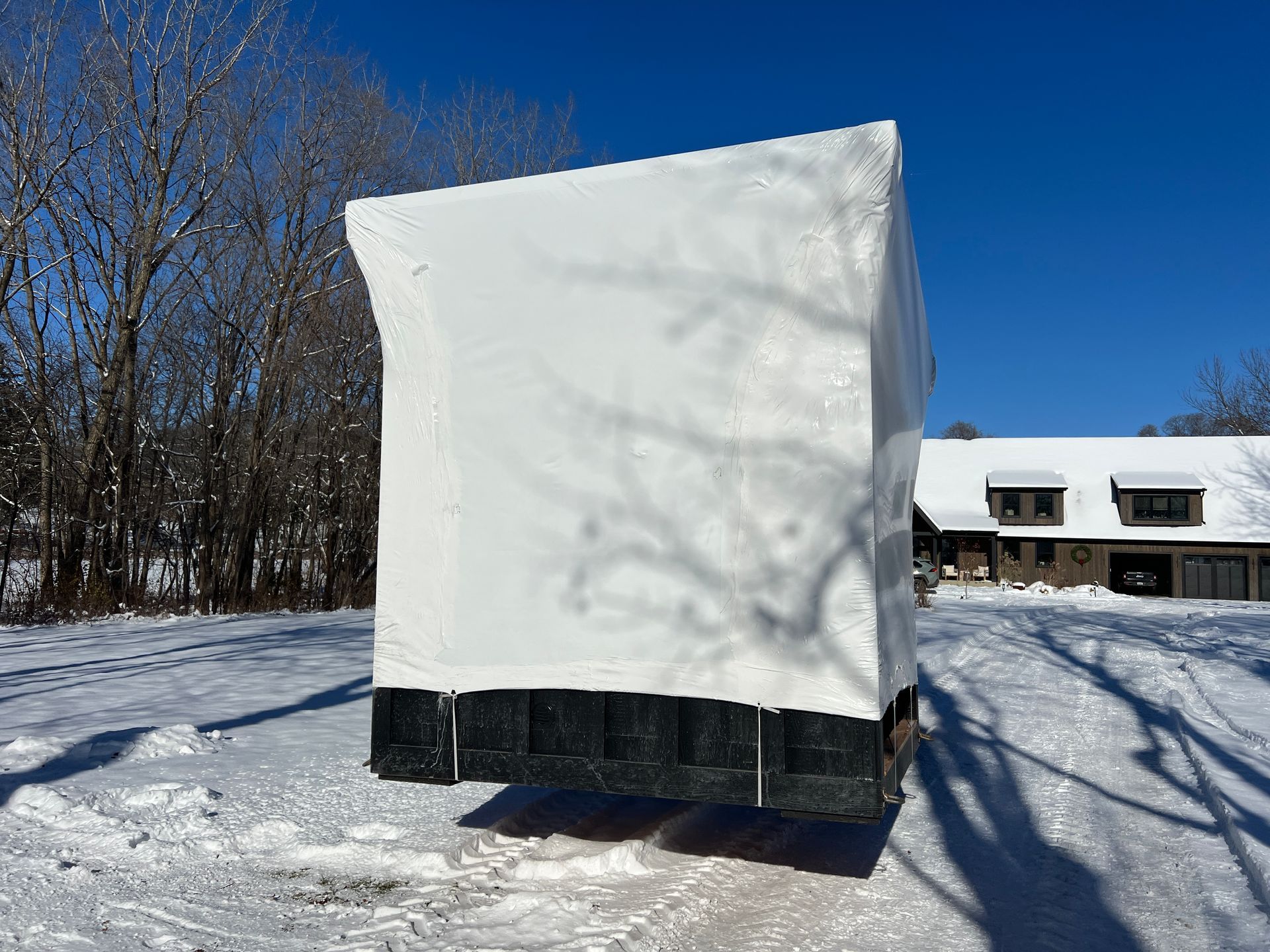A large, white-wrapped block sits suspended over a snow-covered field with a house and trees in the background.