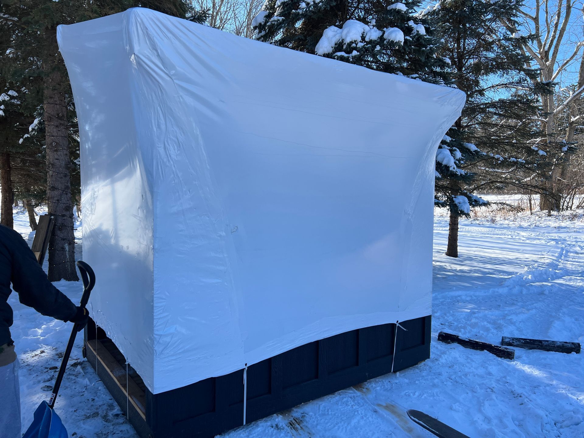 A person with a blue shovel stands next to a large rectangular structure covered in white shrink wrap outdoors in snow.