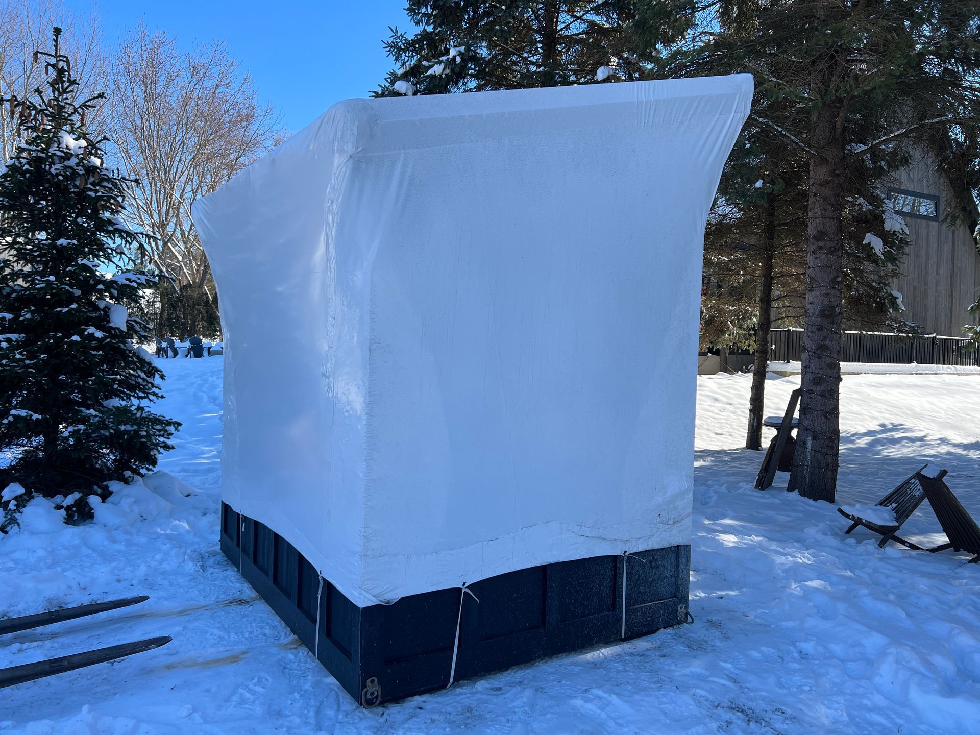 A large structure with a black base is wrapped in white protective shrink wrap, standing in a snowy, wooded area.