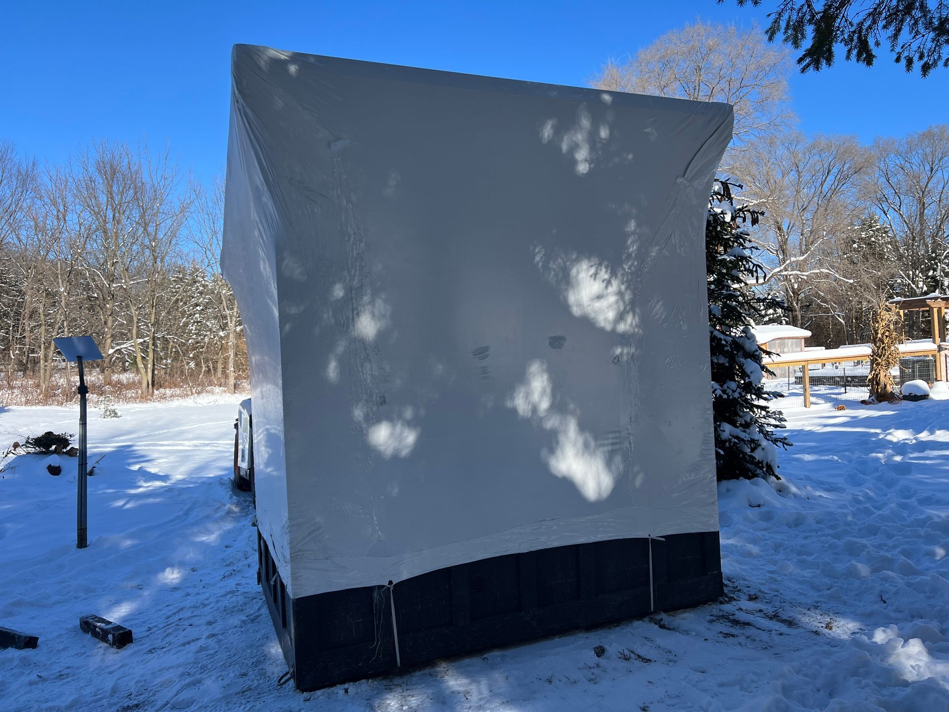 A white, rectangular structure on a base sits in a snowy, wooded area under a clear blue sky.