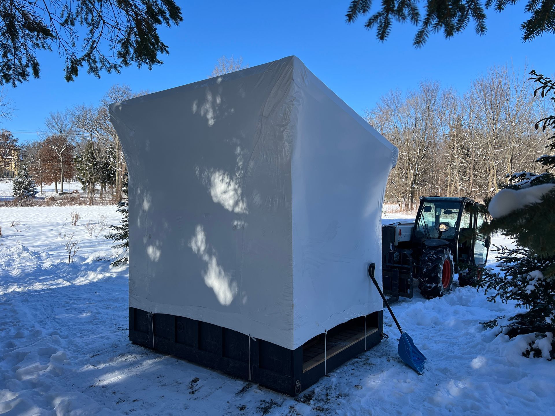 A large, white, square, shrink-wrapped structure sits in a snowy field next to a tractor and a blue snow shovel.