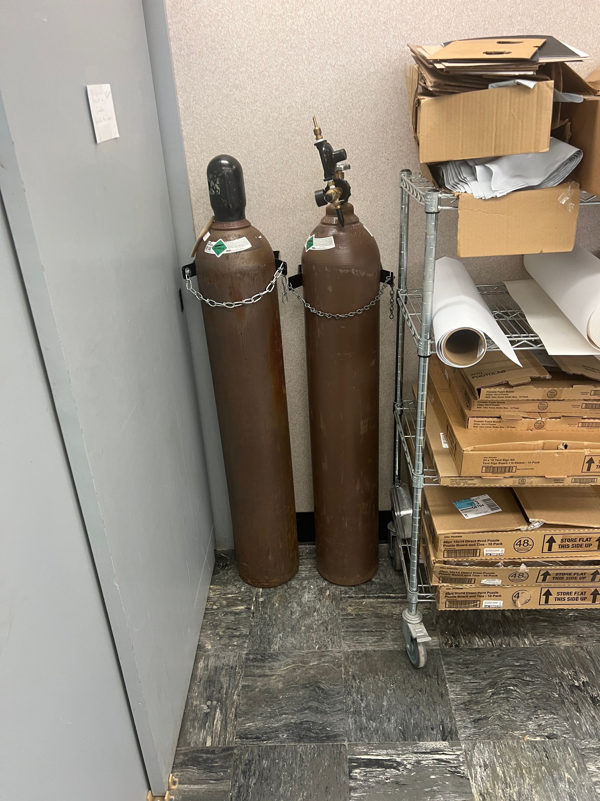 Two brown compressed gas cylinders secured with chains against a wall next to a wire storage rack filled with boxes.
