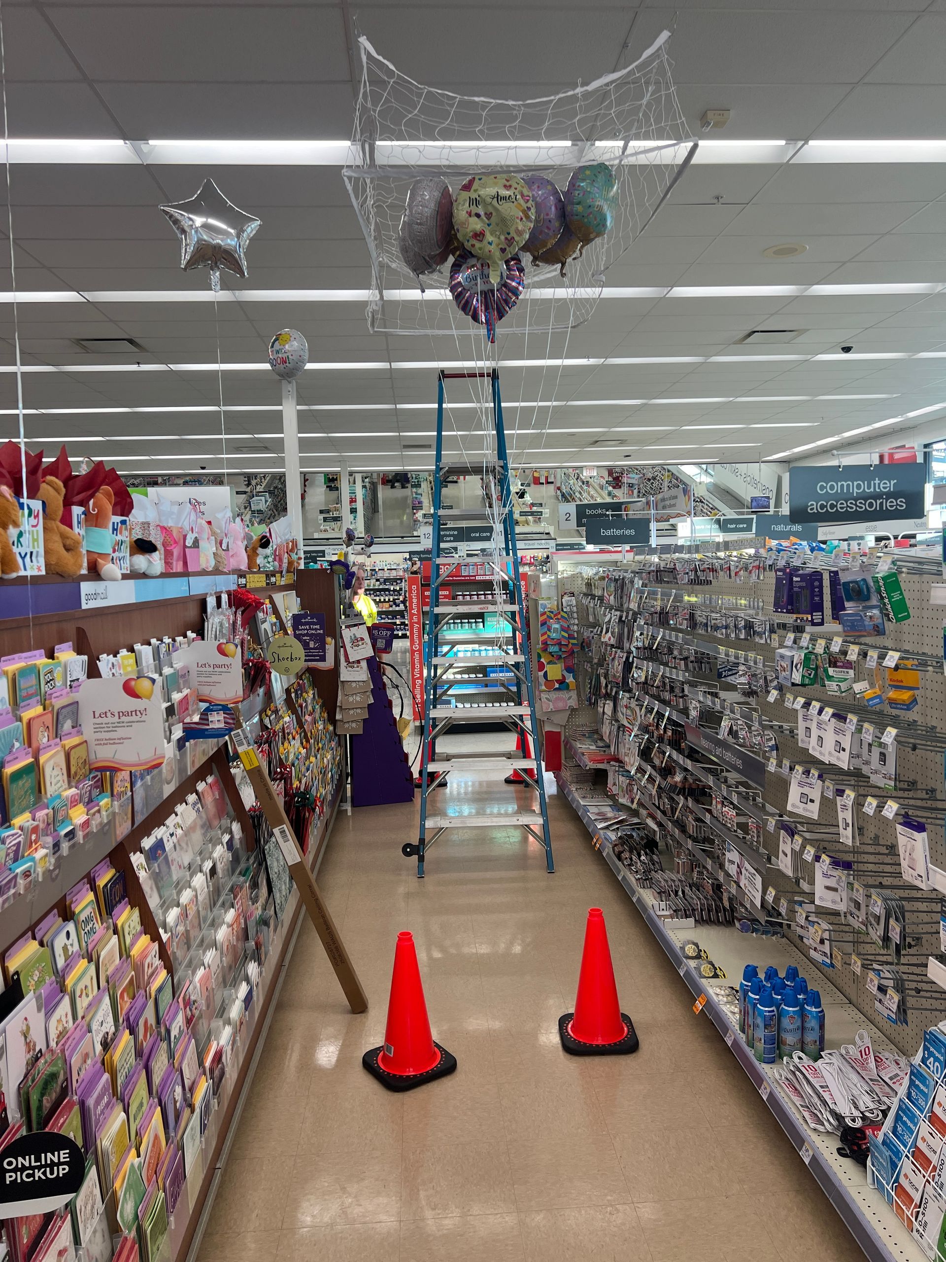 A ladder stands in a retail store aisle beneath balloons caught in a net, with two orange traffic cones on the floor.