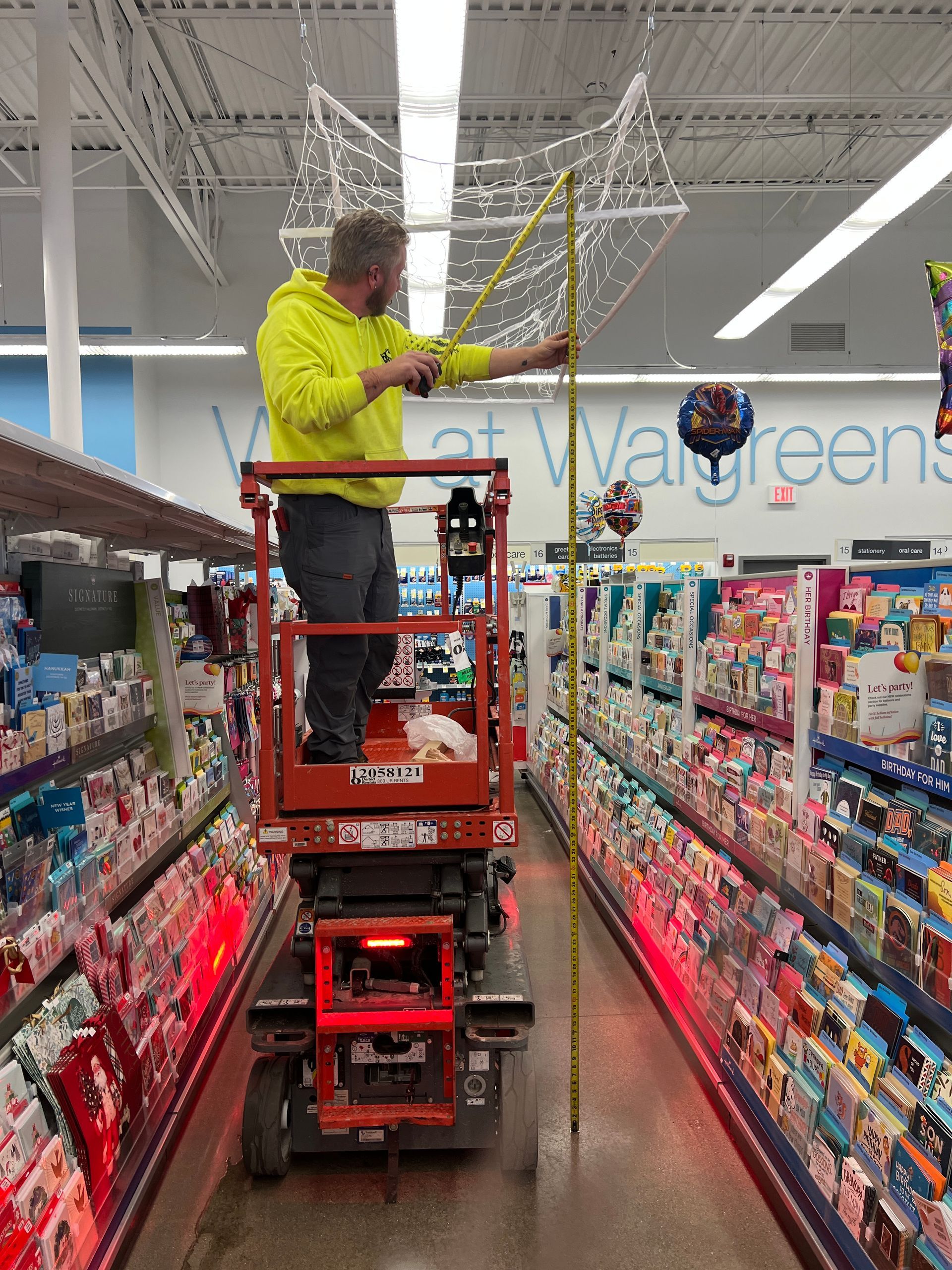 A worker in a neon yellow hoodie on a red scissor lift measures the hanging holiday display in a retail store aisle.