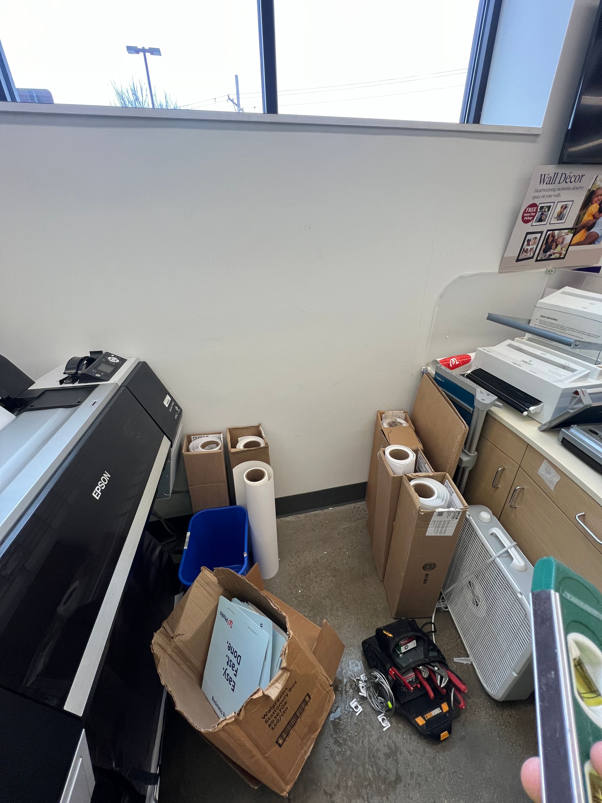 A workspace cluttered with cardboard boxes, paper rolls, a printer, a fan, and a bag on the floor.