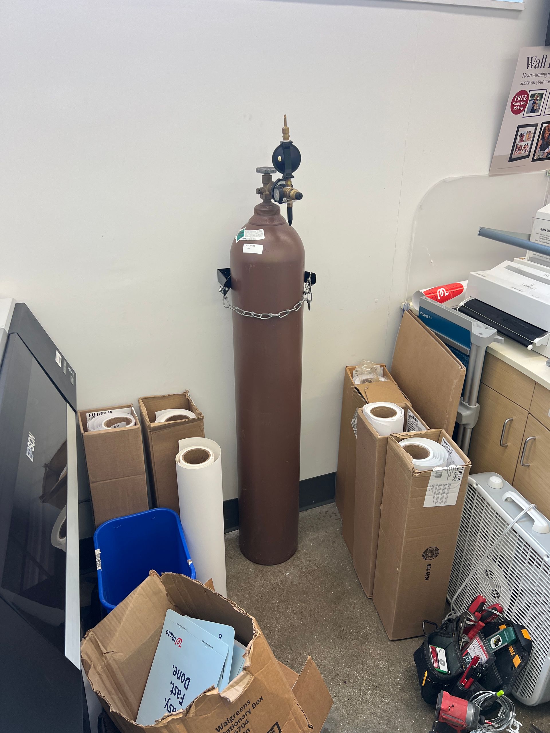 A tall brown gas cylinder secured by a chain to a wall, surrounded by cardboard boxes and office supplies in a room.