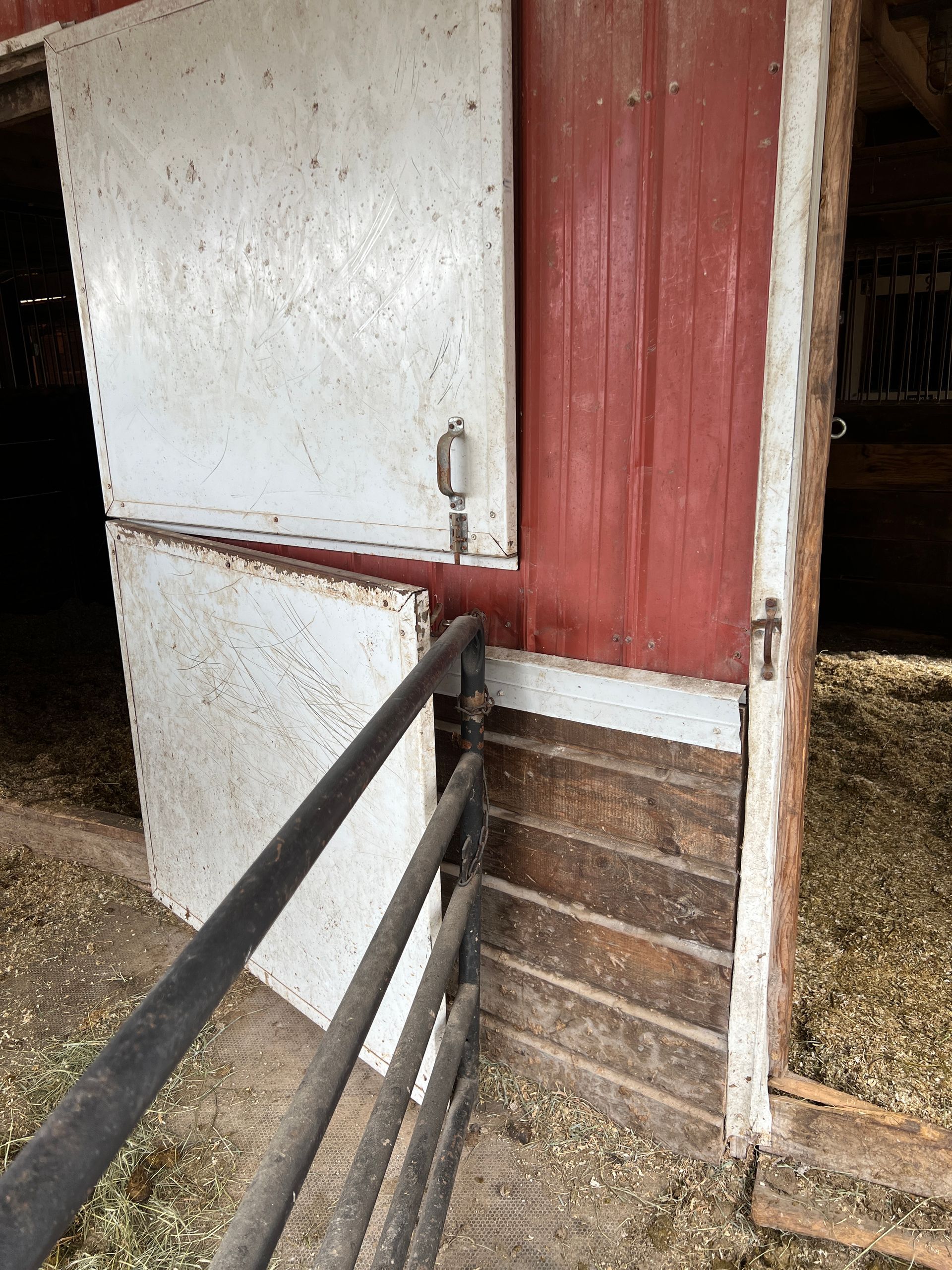 A red and white stall door partially open on a barn, with a metal gate attached and wood-plank flooring covered in bedding.