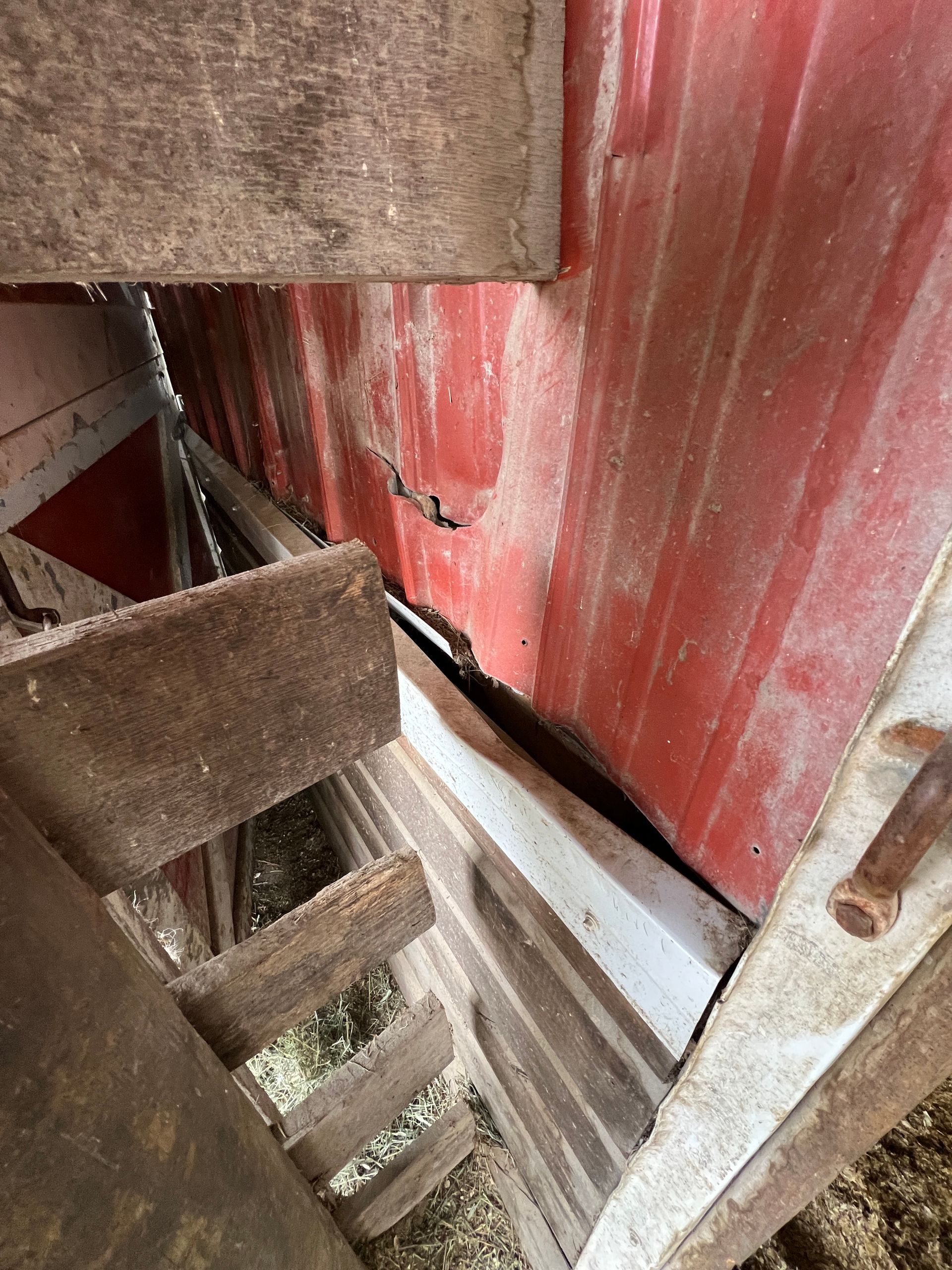 A close-up of a weathered barn interior, showing red metal siding, wooden support beams, and a gap in the structure.