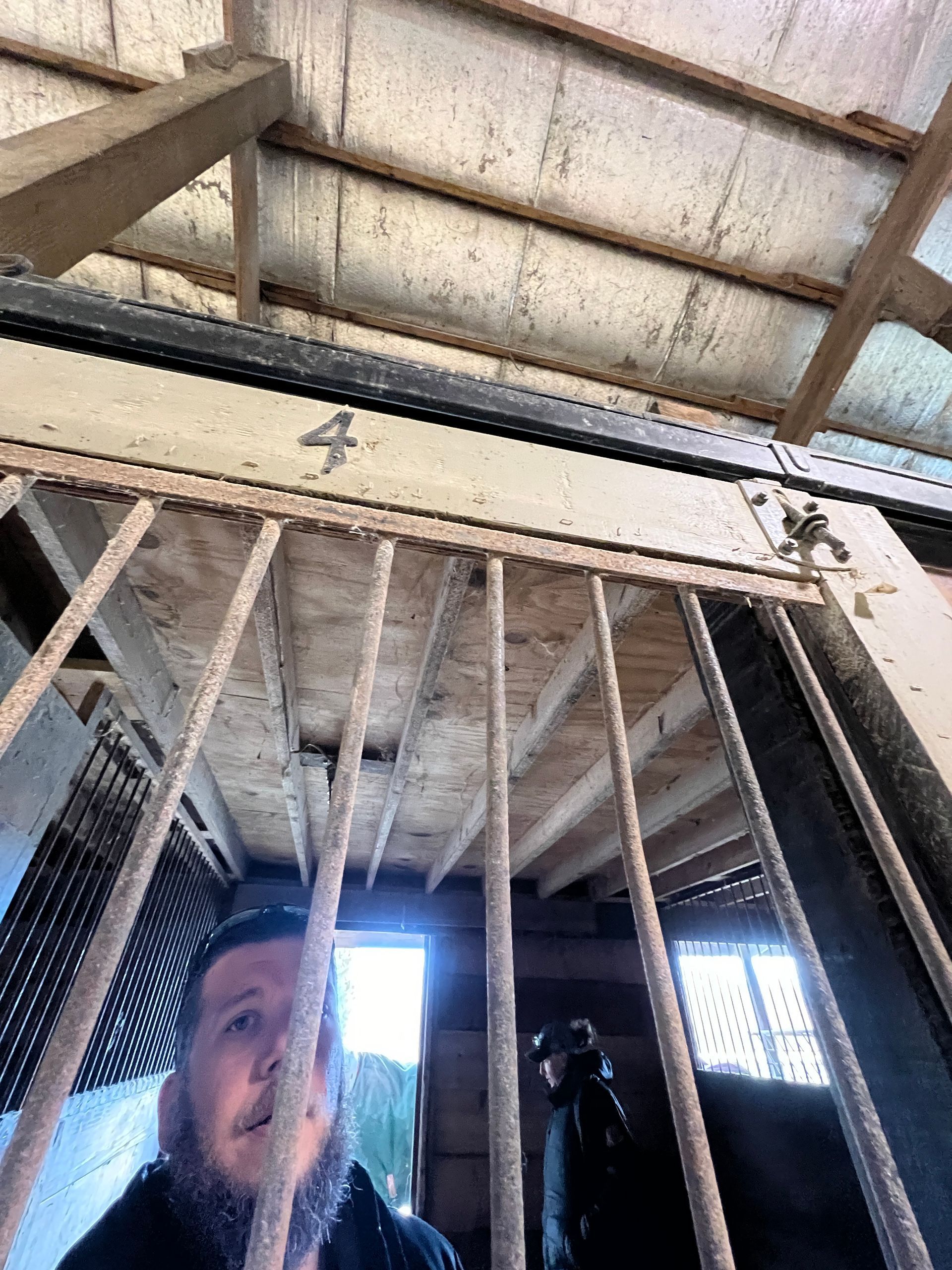 A man with a beard stands behind the bars of a cell labeled with the number 4, with another person visible in the distance.