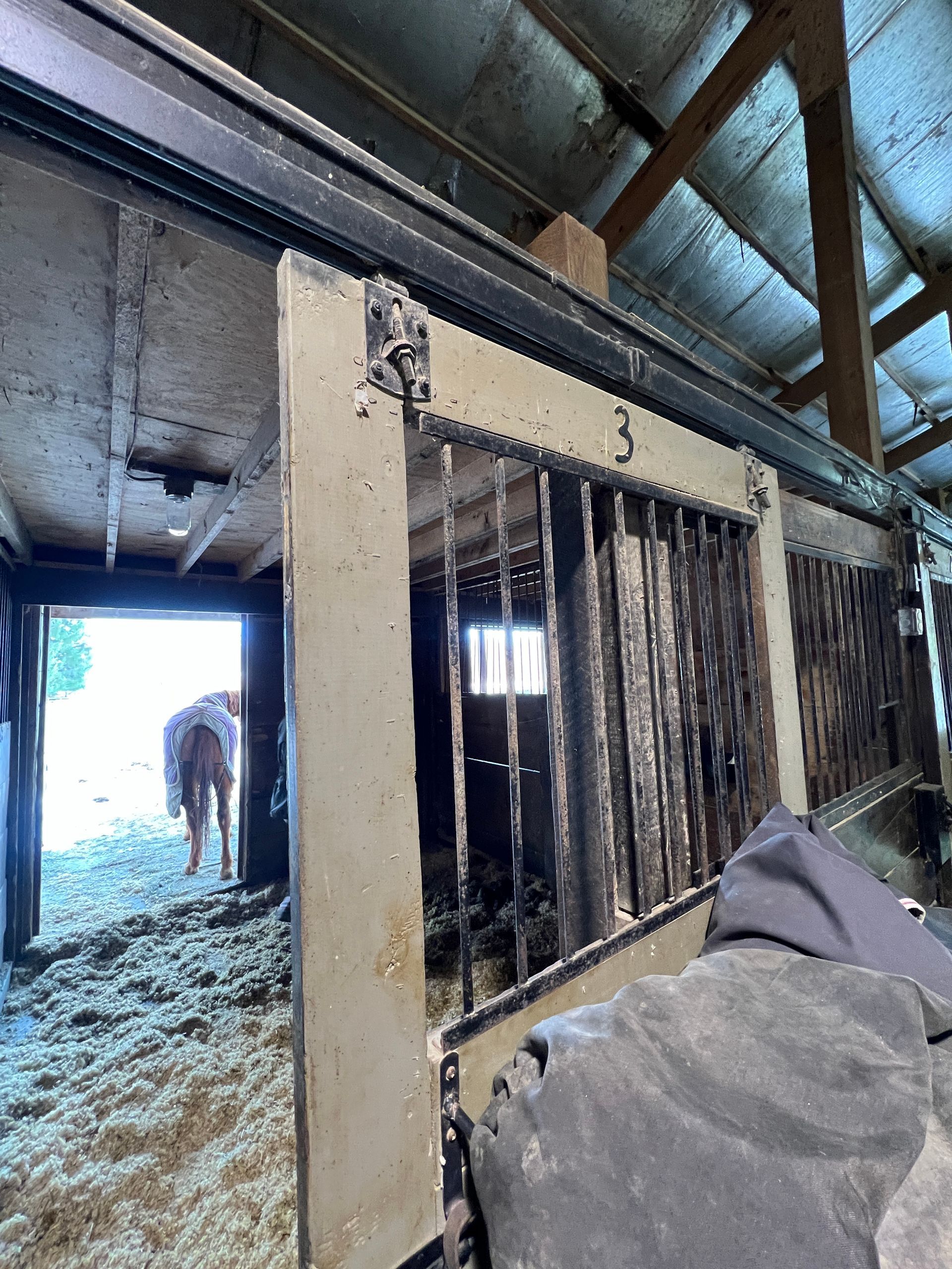 Interior of a horse stable with a numbered stall door in the foreground and a horse visible in a doorway in the distance.