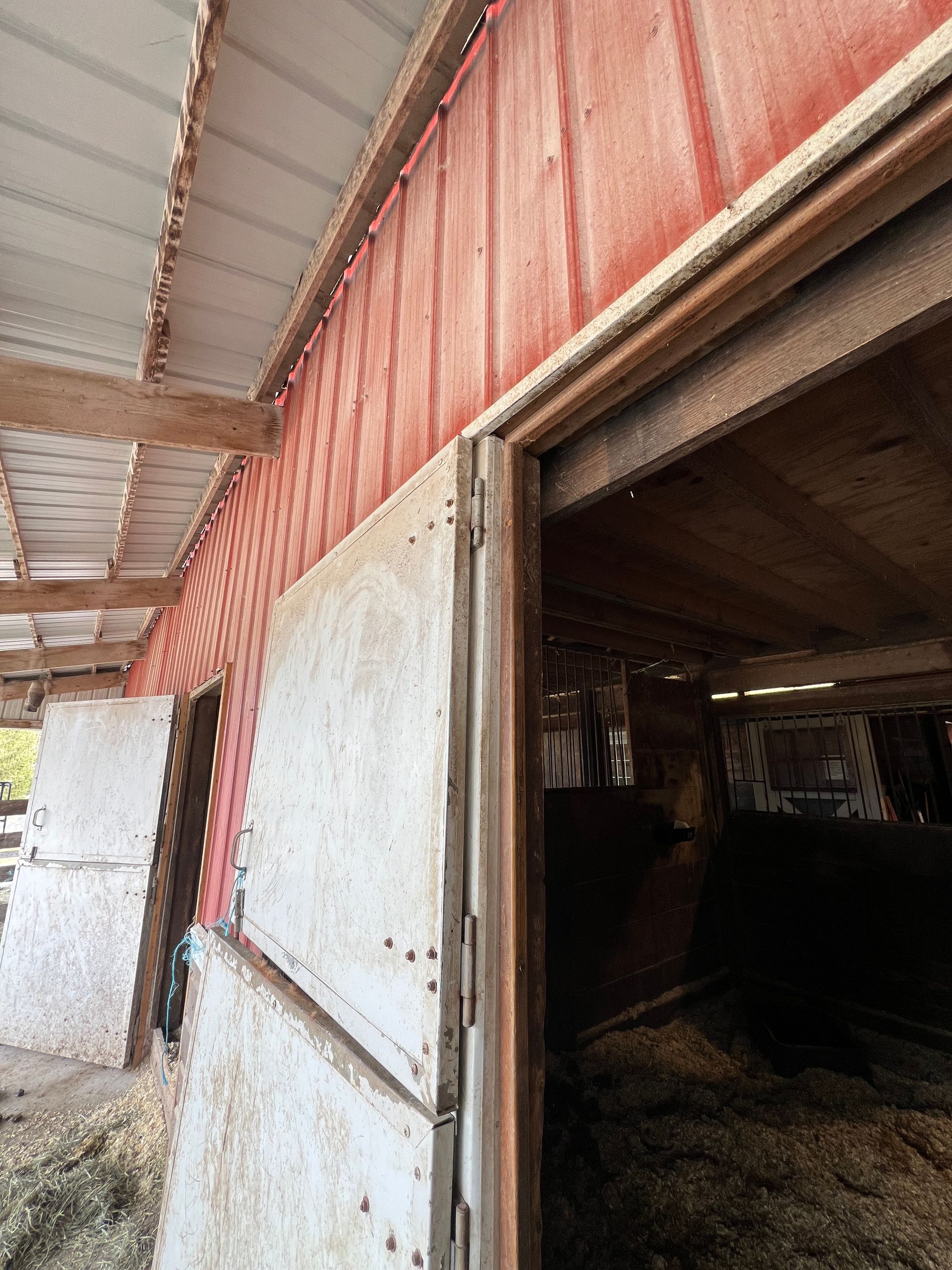 A weathered red barn exterior with white wooden stall doors under a slanted roof, showing a glimpse into a dark stall.