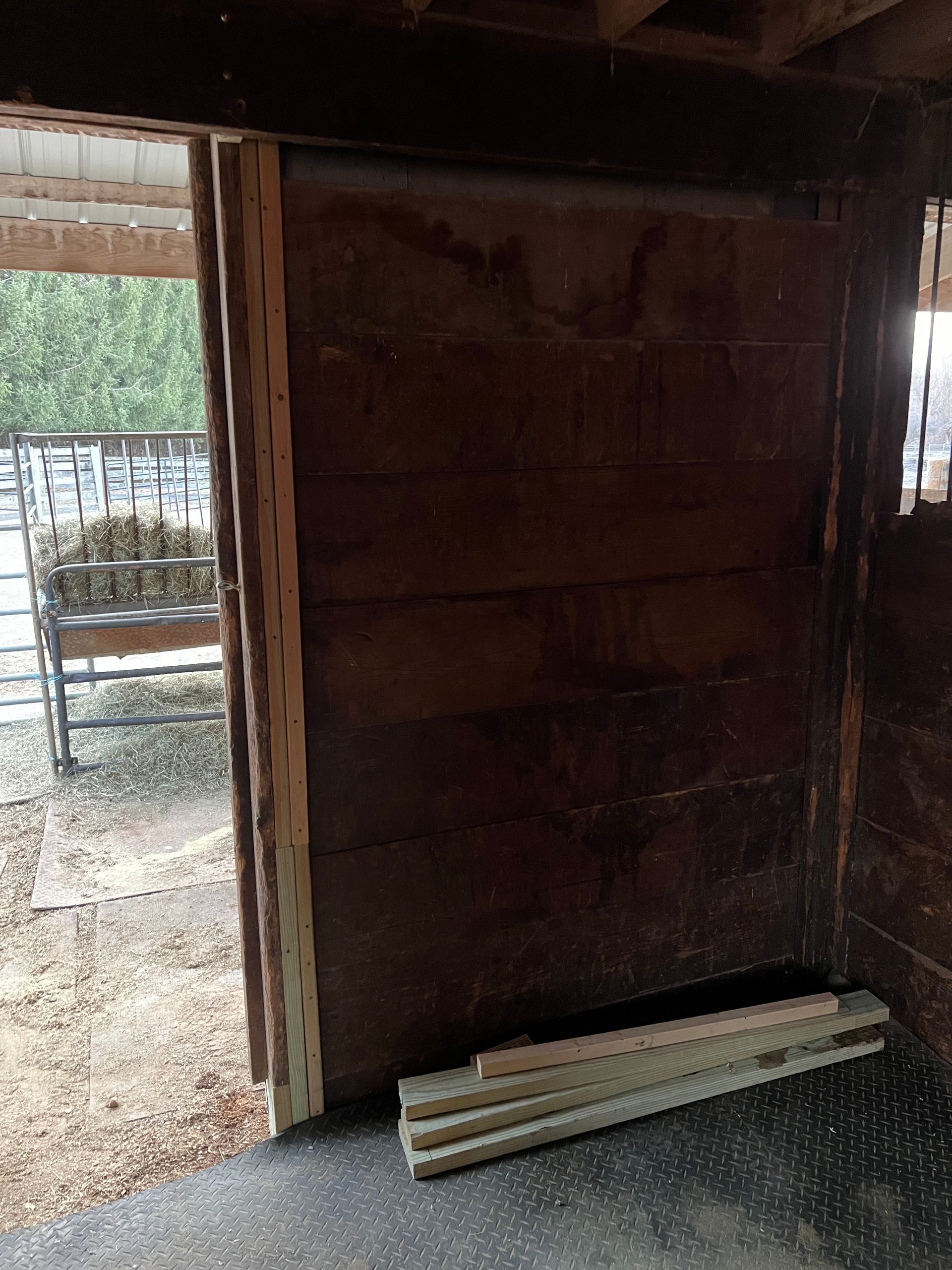 A wooden stall door in a barn with a stack of light-colored replacement boards resting on the floor.