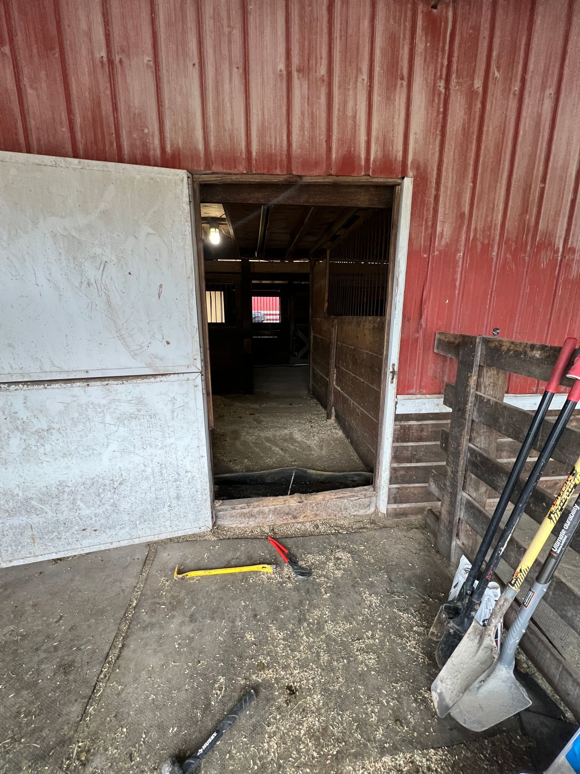 Open door to a dim wooden barn stall with shavings on the floor, red siding, and tools resting on the concrete ground.