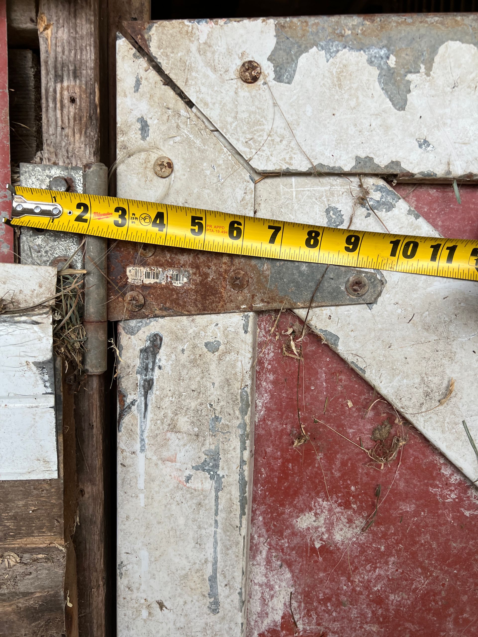 A measuring tape extends 11 inches across a rusted metal hinge attached to a weathered, painted wooden door.