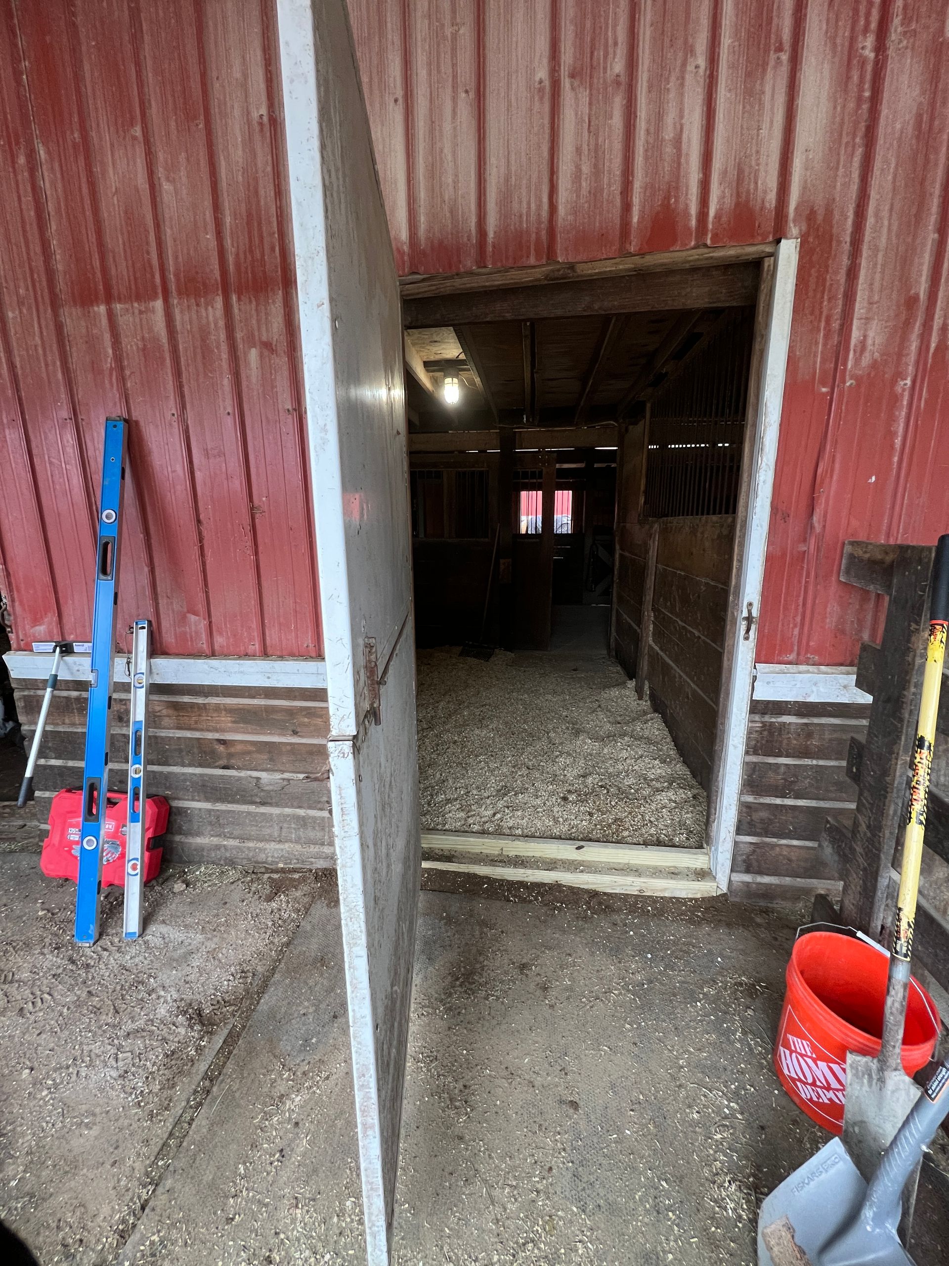 An open door to a barn interior with wood shavings on the floor, red siding, blue skis to the left, and a red bucket.