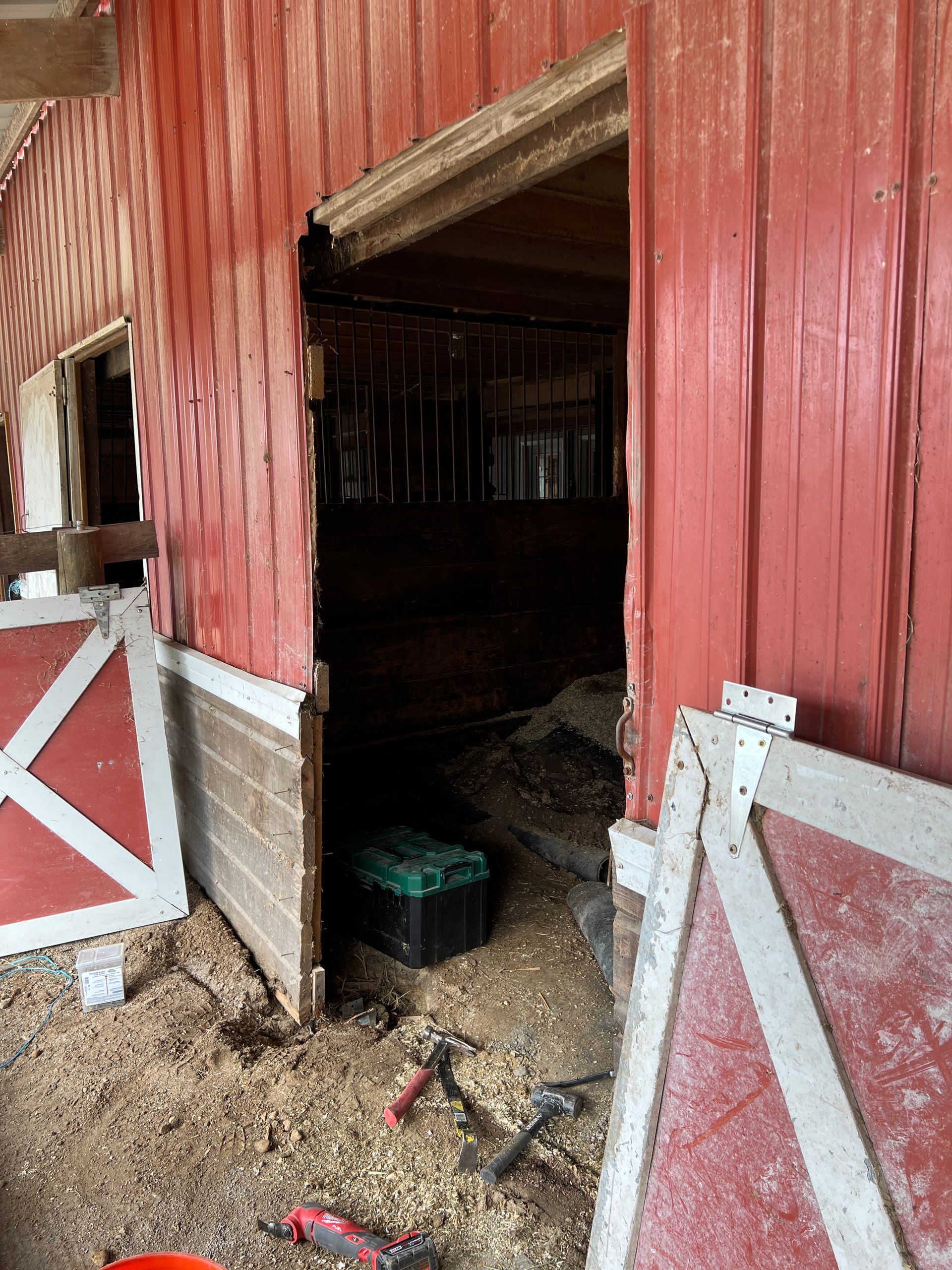 A detached red barn door lies on the ground next to an open doorway of a rustic red barn building.