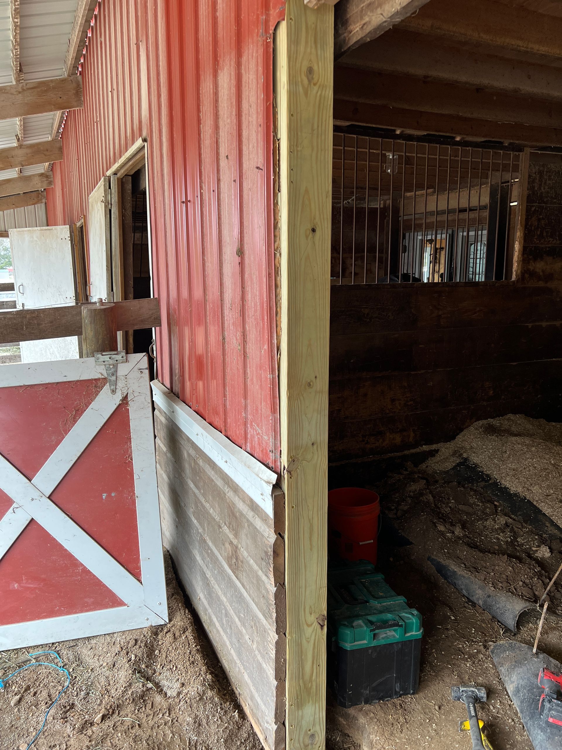 A wooden stall entrance at a red barn, featuring a partial red-and-white door and a fresh light-colored support post.