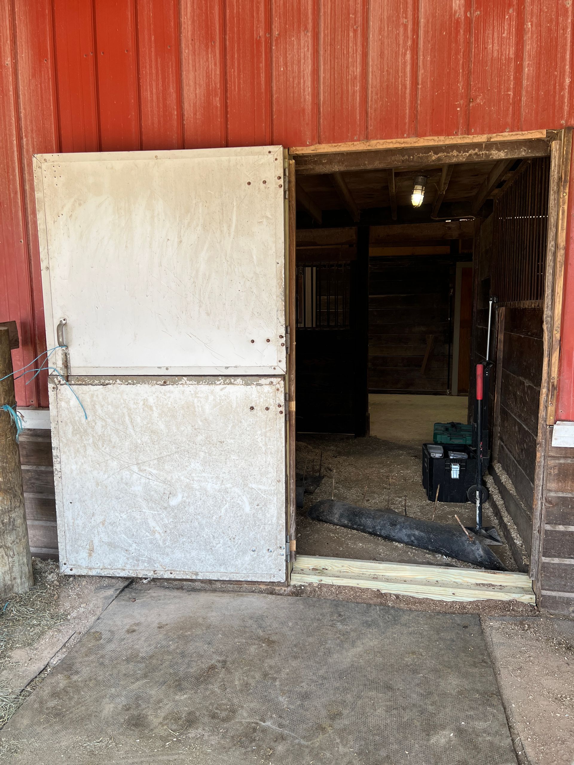 An open barn door showing a dim, interior space with a black box on the floor against a red exterior wall.