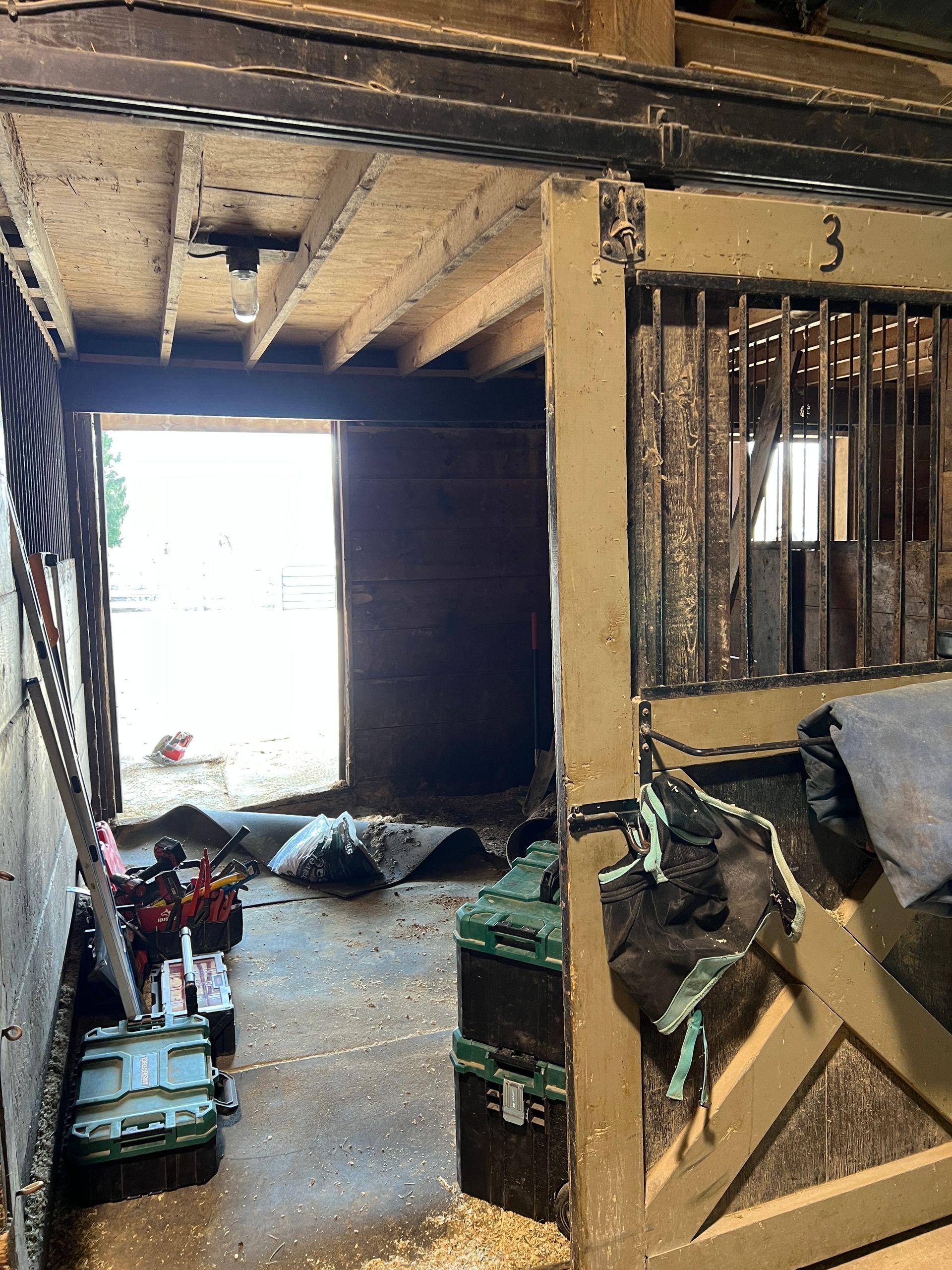 Looking into a stable stall doorway, showing a partially open wooden stall door, tools, and a bright outdoor view.