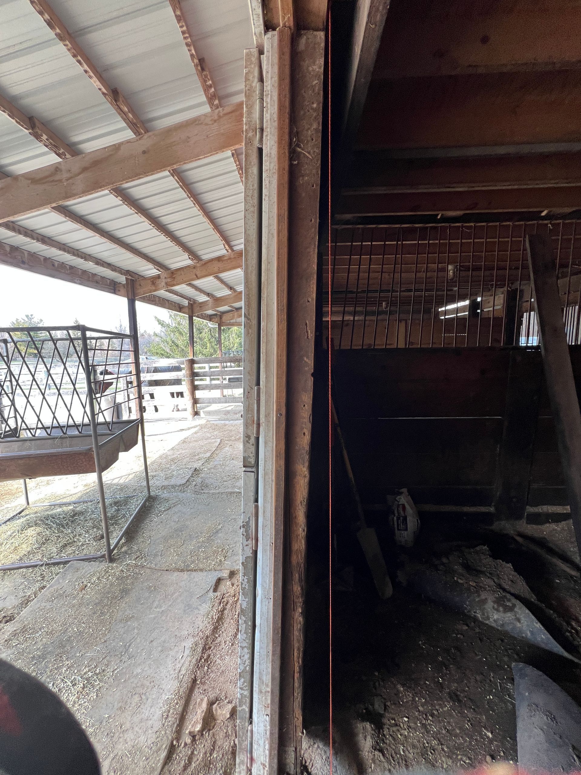 A dark, weathered barn interior seen from an opening, looking out toward a covered outdoor area with a metal animal feeder.