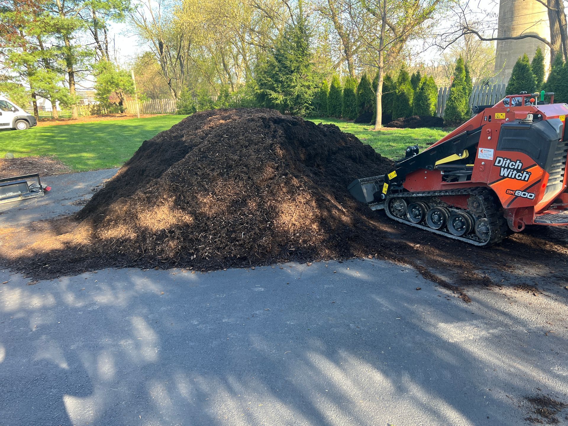 A red skid steer loader parked next to a large pile of dark mulch on an asphalt driveway in a grassy, treed landscape.