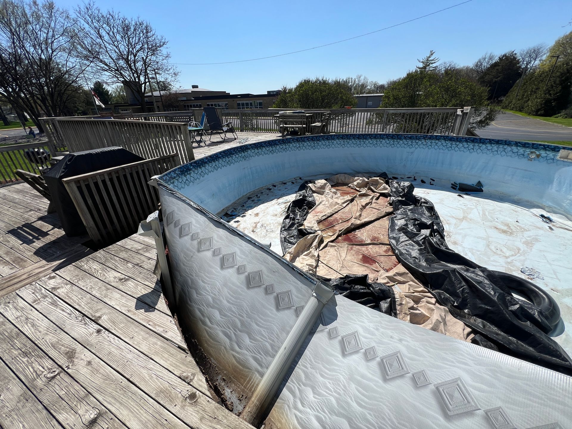 An empty, circular above-ground pool with a weathered white side, on a wooden deck in a yard on a sunny day.