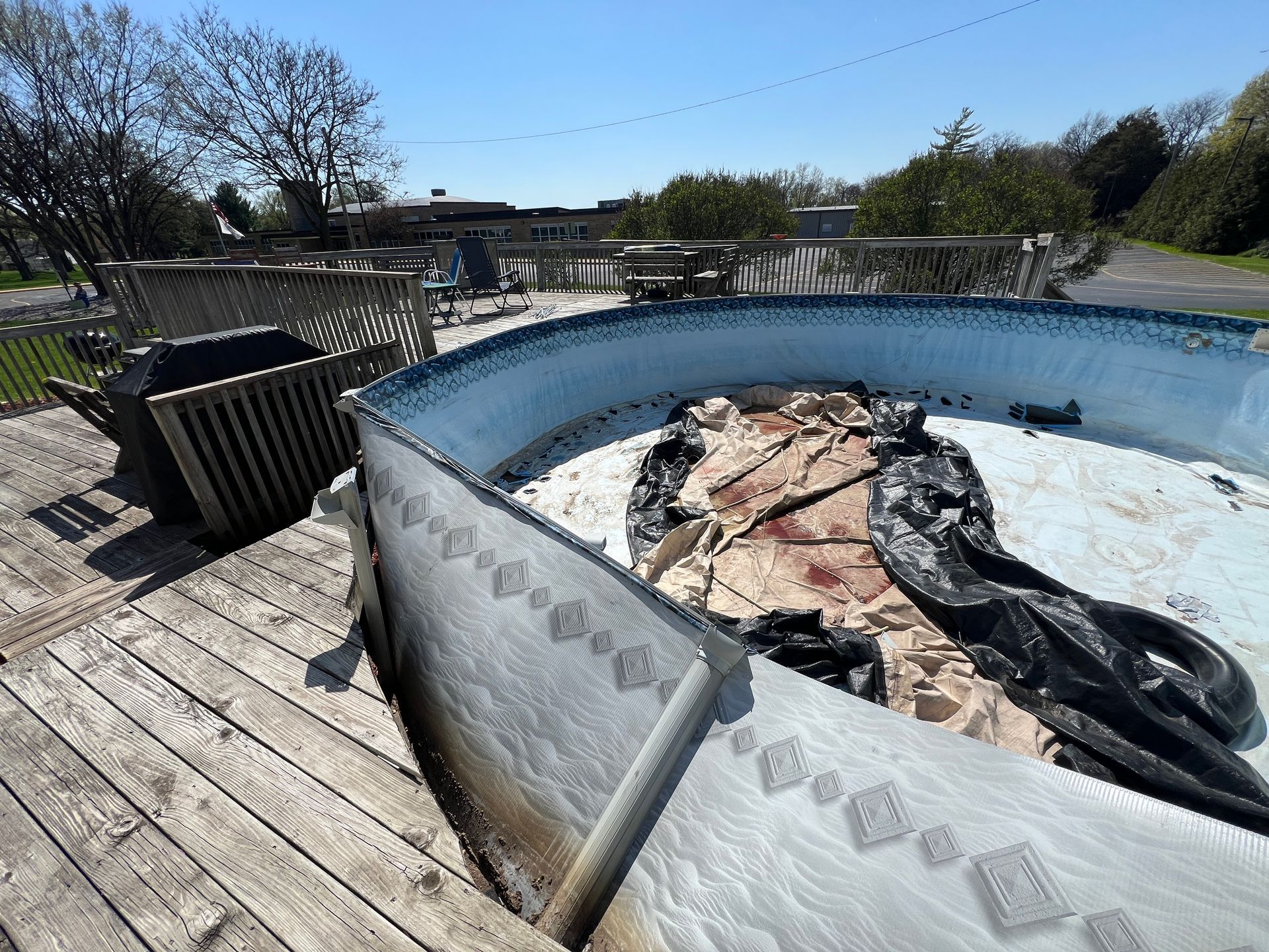 An above-ground pool sits empty on a wooden deck, containing debris and a dark, crumpled pool cover under a clear sky.