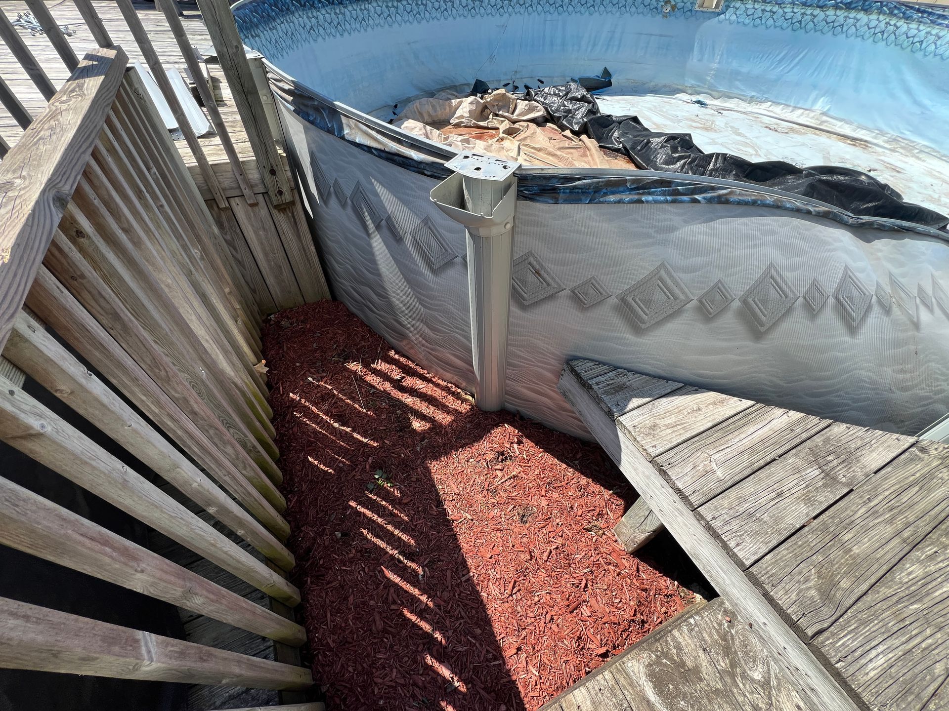 Above-ground pool wall and upright support located between a wooden deck and railing, with red mulch on the ground.