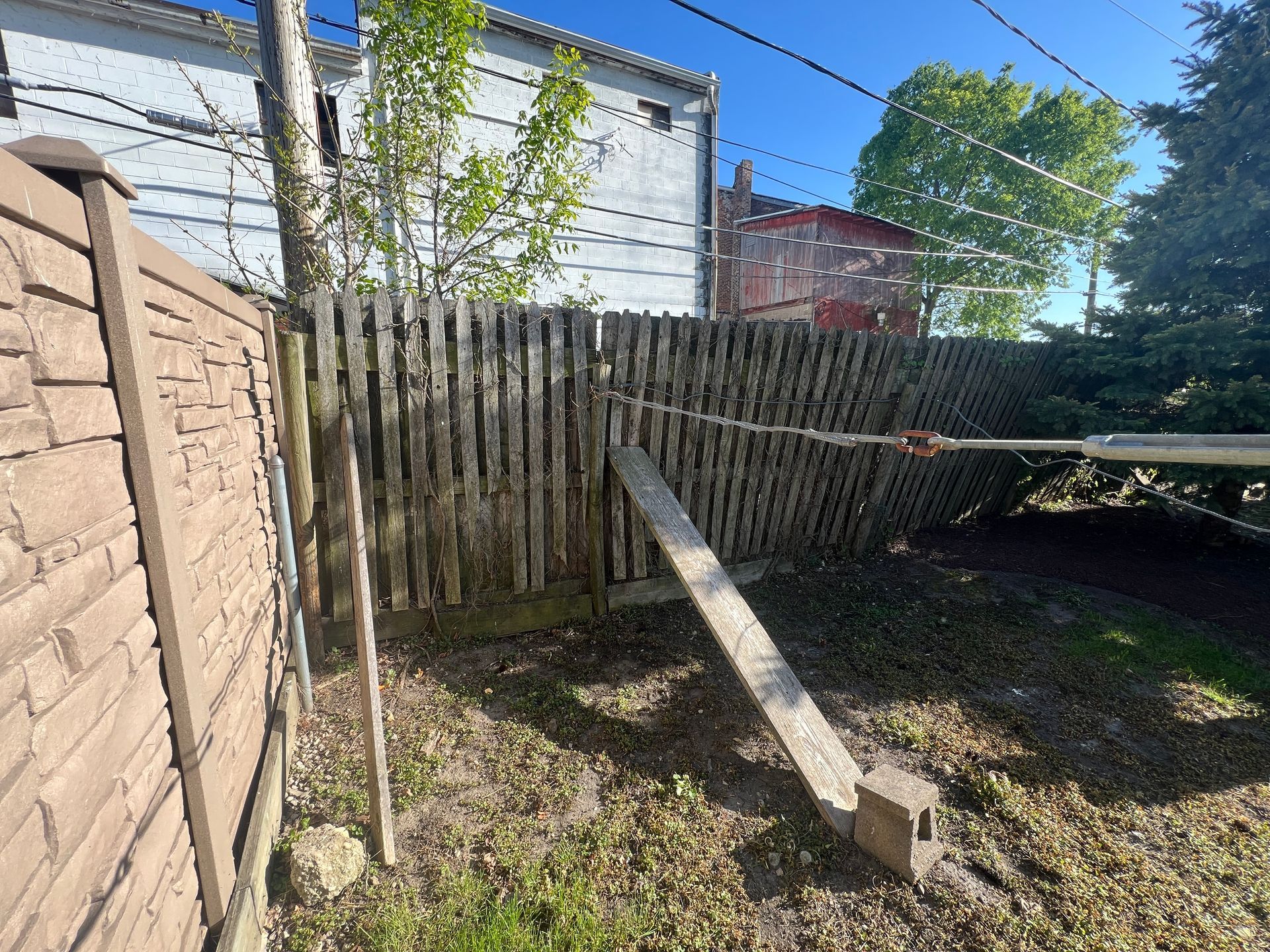 A leaning wooden fence in a backyard is braced by a diagonal wooden board resting on a concrete block.