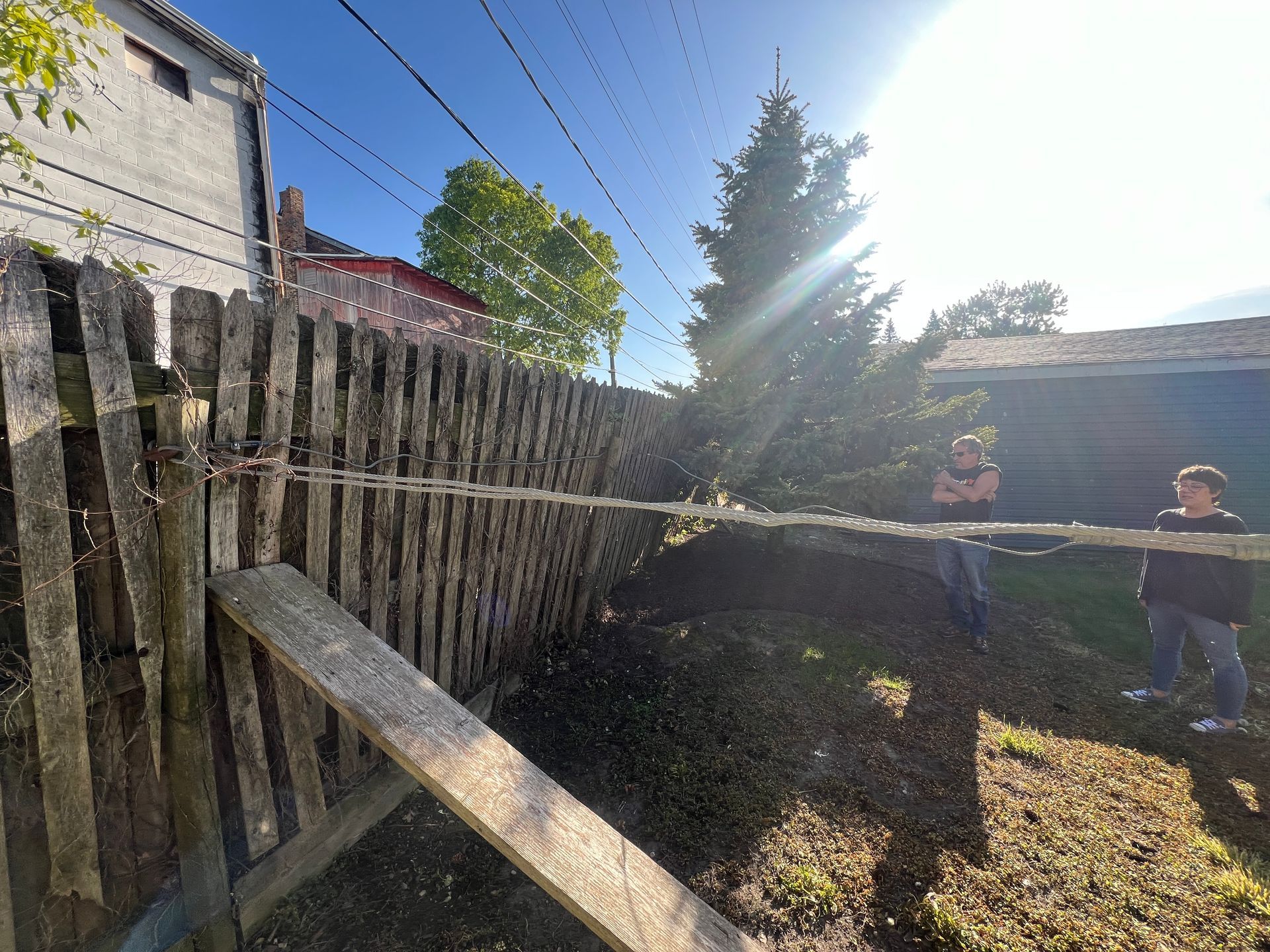 Two people stand in a sunny yard near a damaged, leaning wooden fence and a fallen evergreen tree.