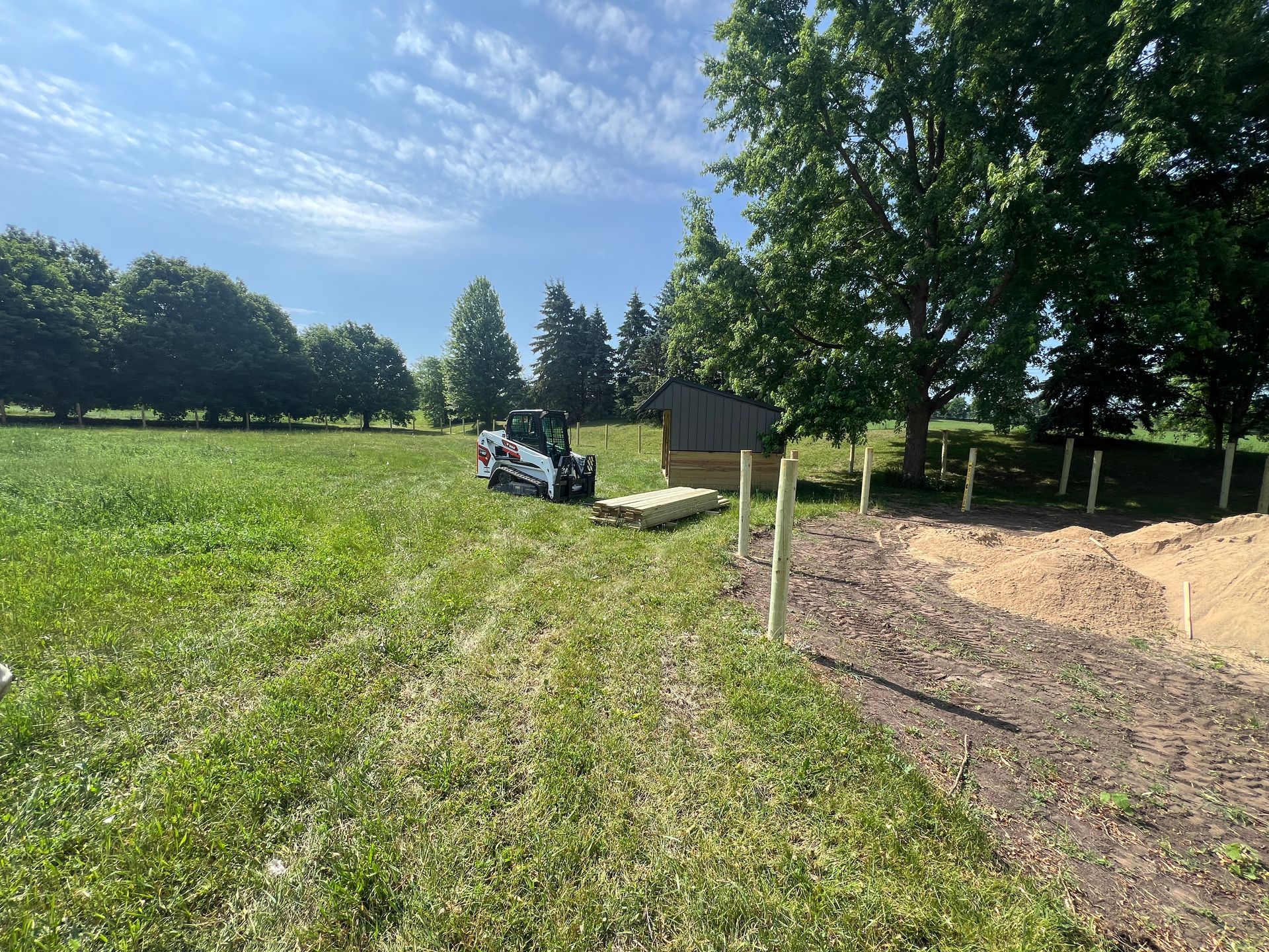 A skid steer sits in a grassy field near a small shed and a line of newly installed wooden fence posts.