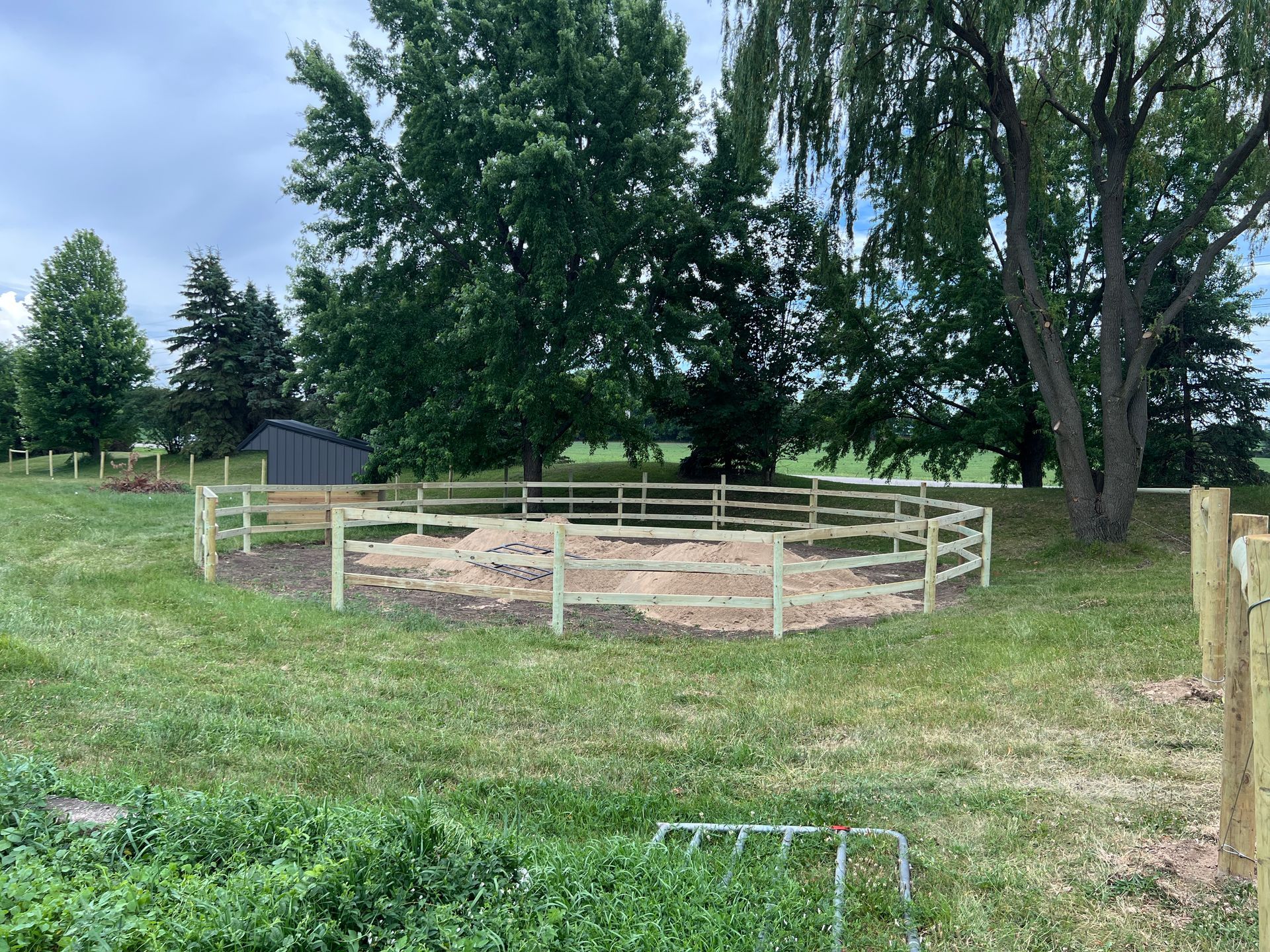 A circular wooden round pen for horses set in a grassy field with trees in the background under a cloudy sky.