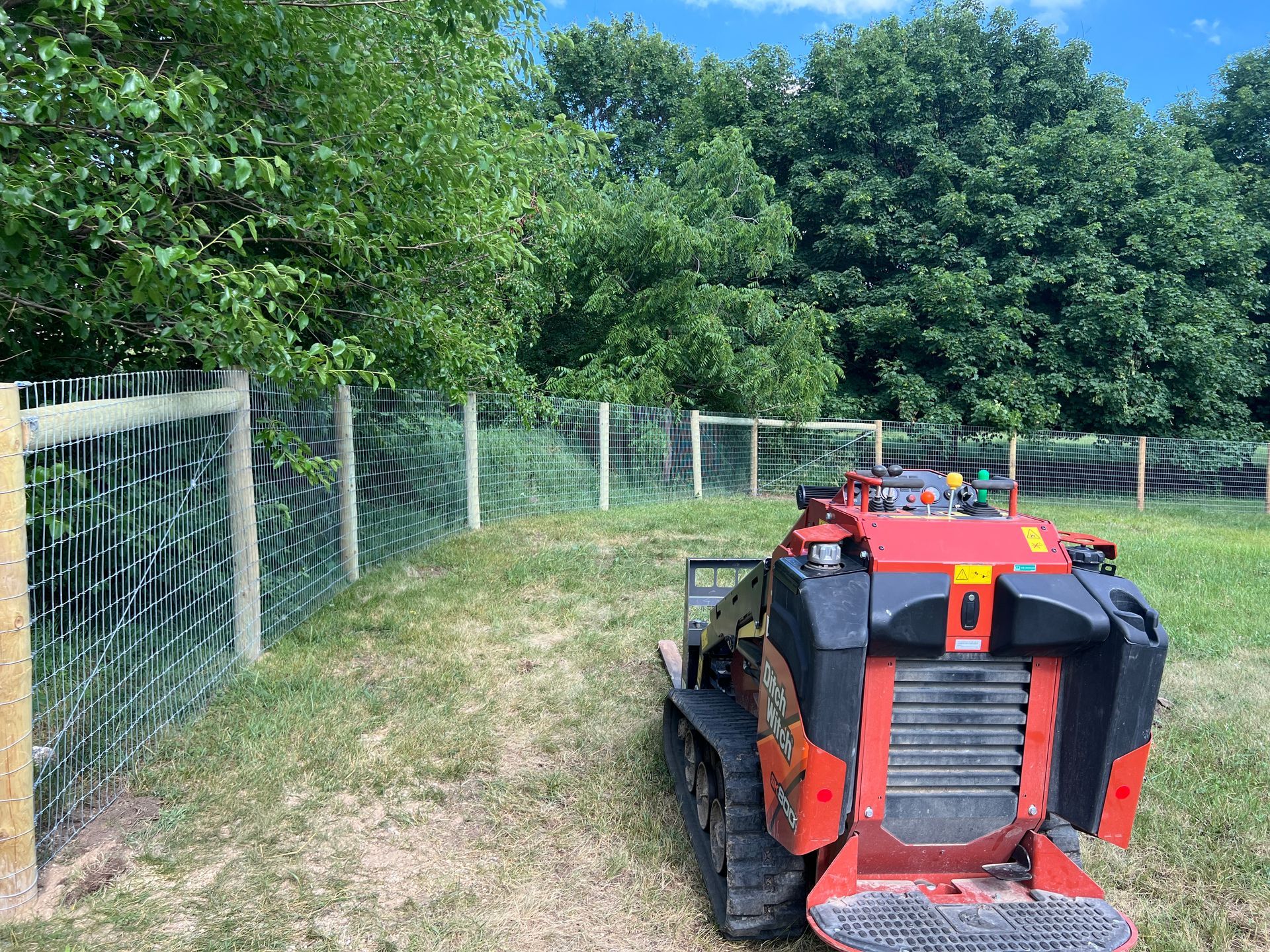 A red tracked skid steer parked on a grassy hill next to a wire fence bordering a line of trees.
