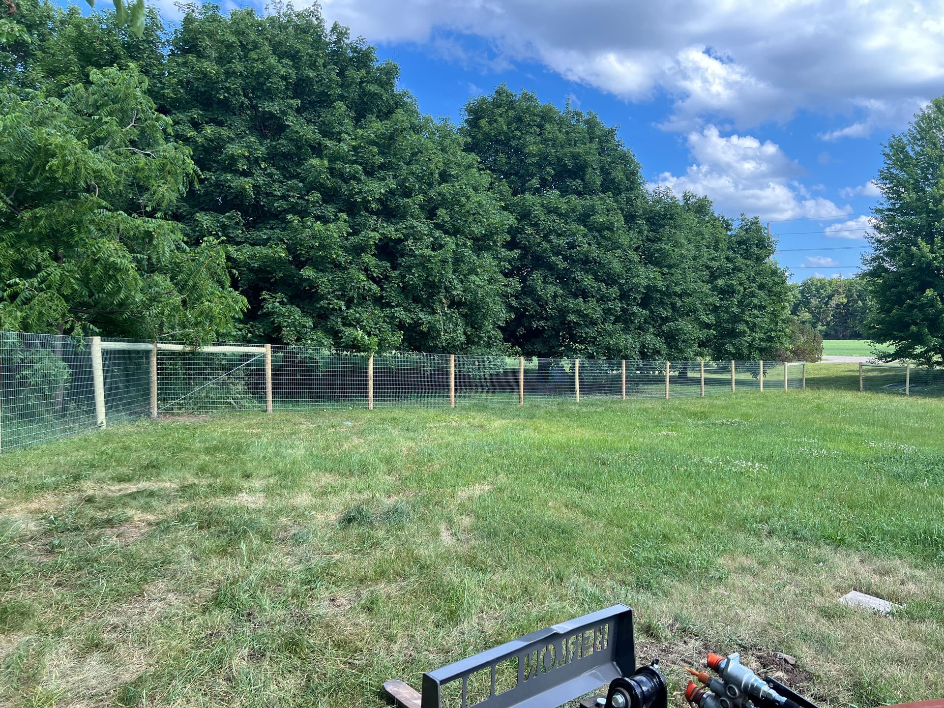 A wire mesh fence stretches across a grassy field in front of a dense line of green trees under a blue, cloudy sky.