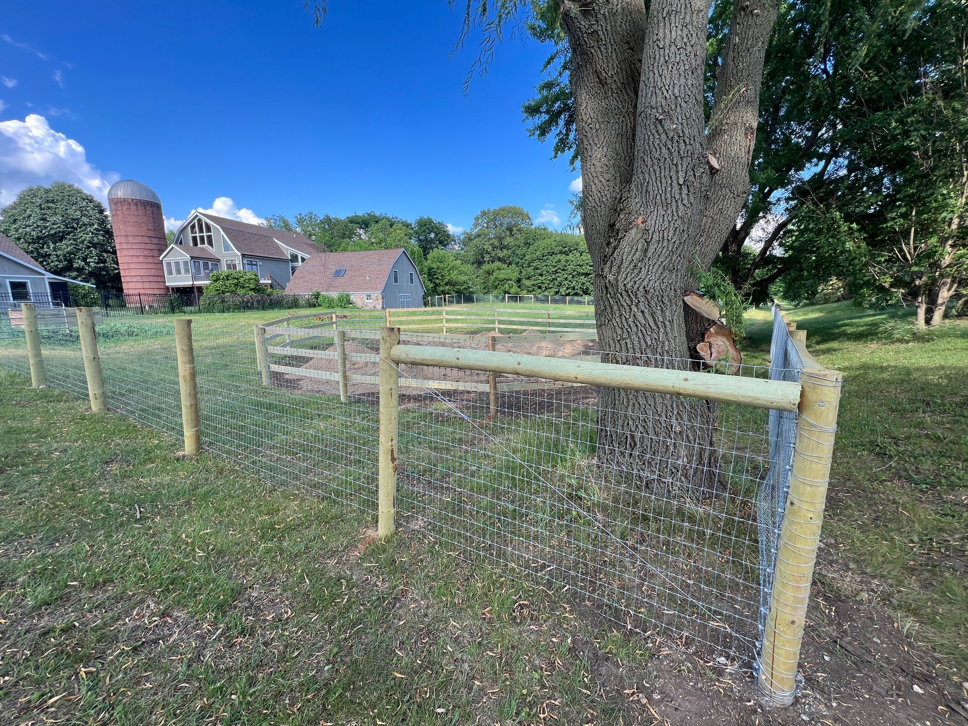 A wooden post-and-wire fence runs through a grassy field with a large tree and a farmhouse with a silo in the background.